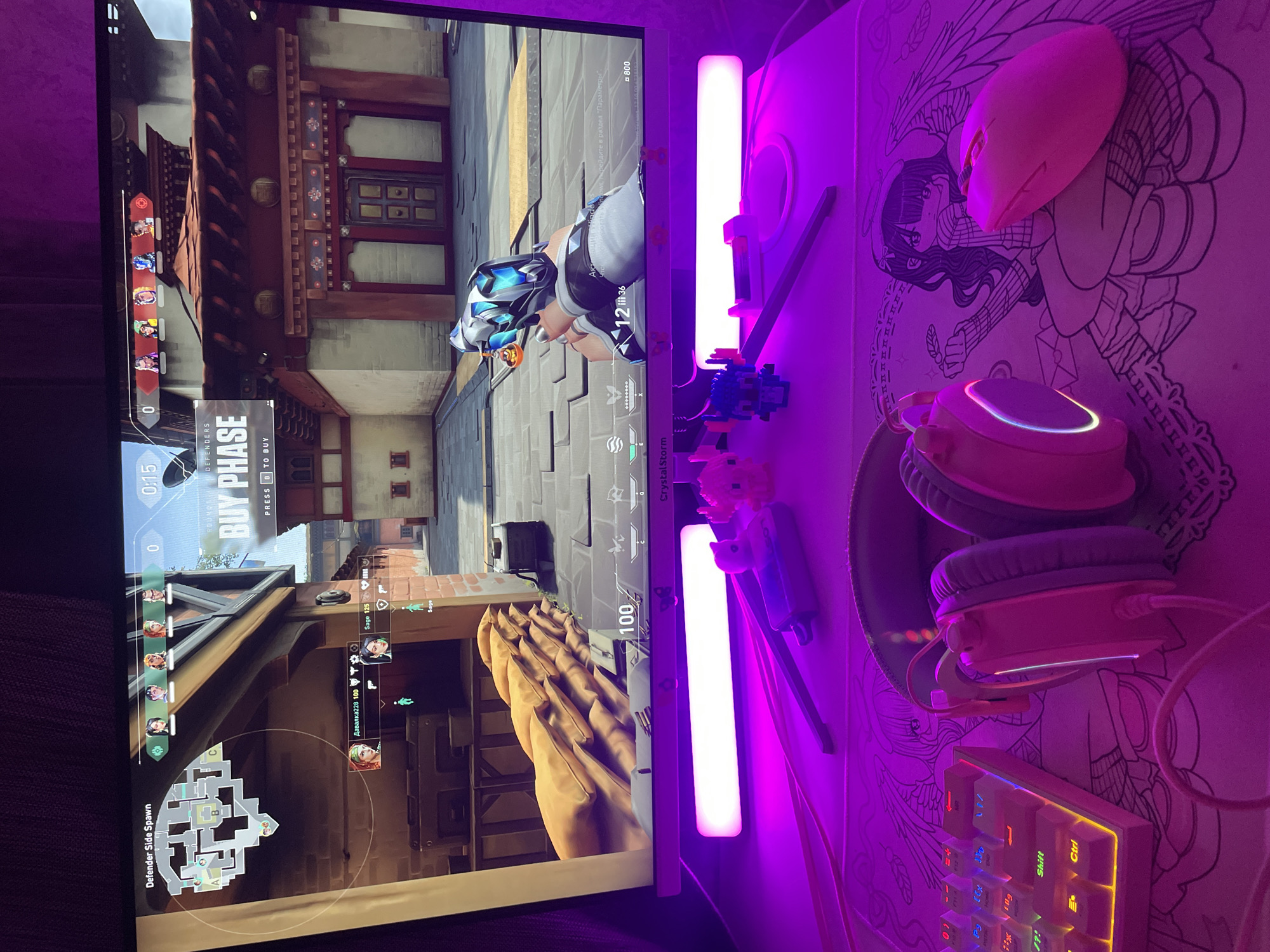 The image captures a vibrant gaming setup, illuminated predominantly by pink and purple RGB lighting, suggesting an evening or nighttime setting in Votkinsk, Russia. A monitor, oriented vertically in the photo, displays a scene from the tactical first-person shooter game Valorant. The in-game view shows a character, clad in blue and white armor with a distinctive helmet, in what appears to be an urban map with traditional East Asian architectural elements, including red-tiled roofs and decorative doors. The game is currently in the "BUY PHASE" for "DEFENDERS," with a 0:15 countdown timer and an active minimap showing the "Defender Side Spawn." Visible on-screen text includes player names and scores such as "Давалка228 100" and "Sage 125," along with game currency "■800" and a Russian system message: "ows, перейдите в раздел "Параметры"."
Adjacent to the monitor, the desk is equipped with a matching set of gaming peripherals. Two vertical LED light bars cast a bright pink and purple glow across the workspace. A pink wireless gaming mouse is positioned on a large desk mat that features an intricate, anime-style monochrome illustration of a winged female figure. Below the mouse, a pair of pink gaming headphones with glowing RGB rings rests on a stand. In the foreground, a compact mechanical keyboard with vibrant RGB backlighting is visible, displaying keys like "Shift" and "Ctrl." The meticulous setup highlights an engaging and personalized gaming environment.
