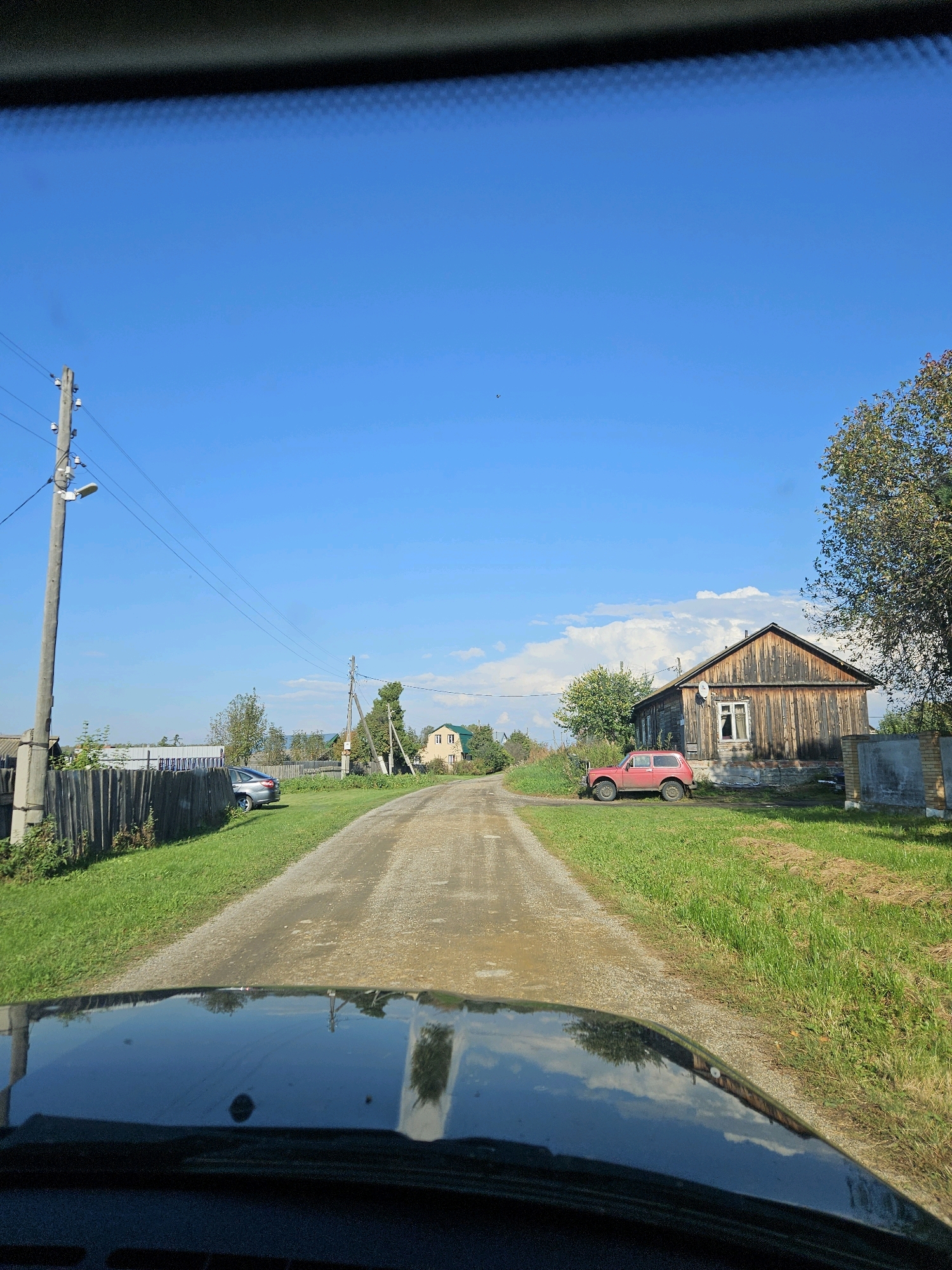 The image shows a dirt road in the village of Sipavskoye, Russia. On the left side of the road, there is a wooden fence, a power pole with streetlights and wires, and a parked car. Further down the road, there are some houses and trees. On the right side of the road, there is a wooden house, a red car, and a large tree. The sky is clear and blue with some clouds. The scene appears to be a sunny day.