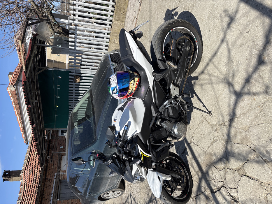 The scene depicts a white and black Kawasaki Z650 motorcycle parked on a cracked concrete surface in Odrintsi, Bulgaria, under a clear blue sky. The motorcycle is positioned facing left, resting on its kickstand. A white helmet, featuring an aggressive shark or joker face design with a blue visor and sharp red-and-white teeth graphics, rests on the motorcycle's black passenger seat. The rear wheel exhibits white, green, and red rim tape. Behind the motorcycle, a dark-colored, dusty Skoda sedan is parked. In the background, two residential-style buildings are visible. The structure on the left is a brick building with a red tiled roof and a chimney. To the right, another building with a lighter-colored facade, also topped with a red tiled roof, is partially covered by a corrugated metal canopy. A dark green tarp or fabric hangs beneath this canopy, equipped with clothesline clips. A white picket fence runs along the right side of the background. A leafless tree stands behind the fence, casting shadows across the foreground. No individuals are present.