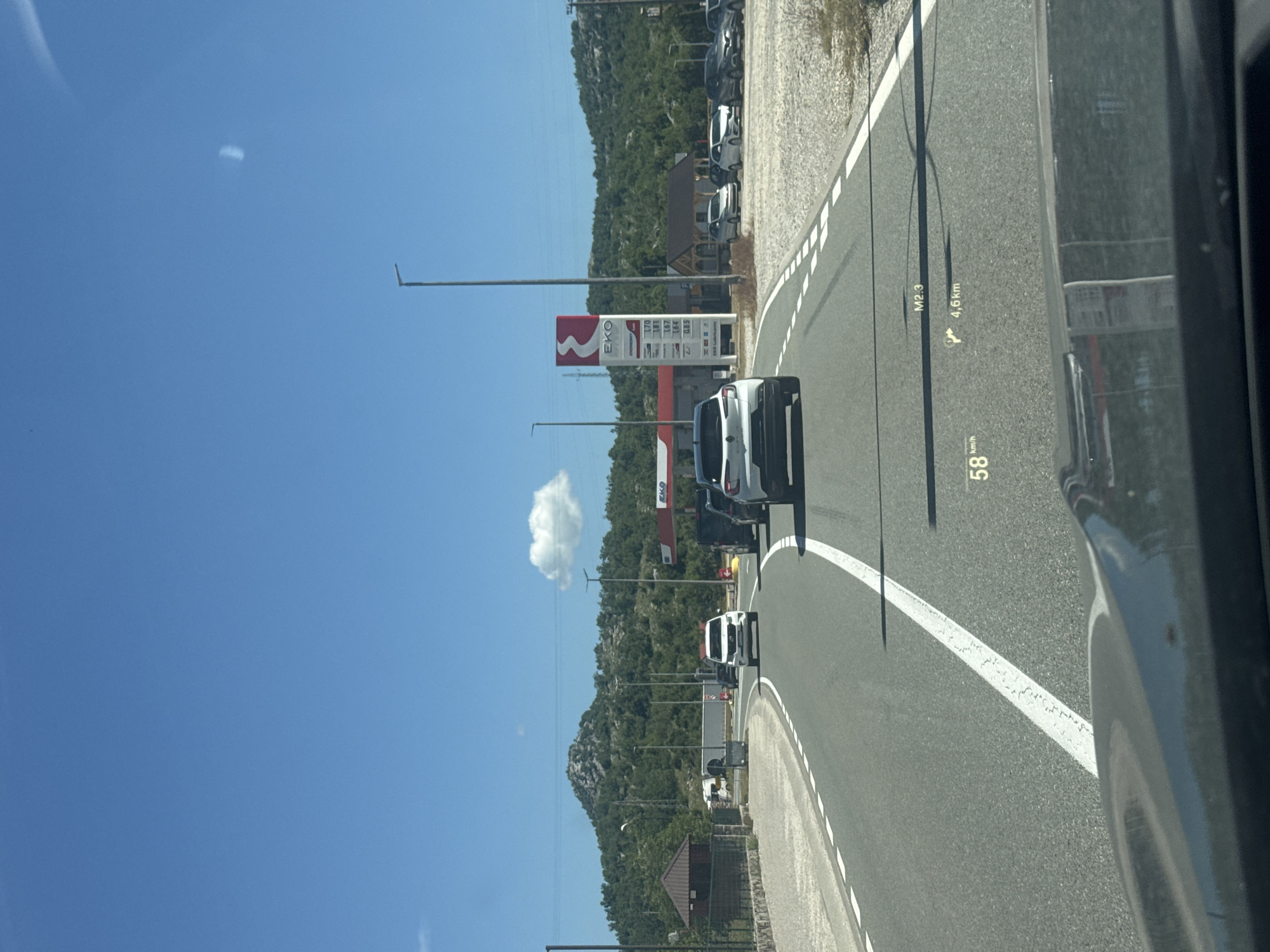 The image captures a view from a vehicle's dashboard on a sunny day, showing a road leading towards a gas station with the EKO brand. The road is asphalt, marked with white lines. Several cars are visible on the road, including a white SUV in the foreground and a white sedan further ahead. The gas station features a tall red and white sign displaying fuel prices. To the left of the road, there's a small brown building with a stone base and a wooden roof, possibly a kiosk or rest stop. In the background, there are green, rocky hills under a clear blue sky with a few scattered clouds. A head-up display (HUD) on the vehicle's windshield shows the current speed as 58 km/h, a distance of 4.6 km, and an indicator for M2.3. The image appears to be taken on the highway near Očinići, Montenegro, given the context of the EKO gas station and the surrounding landscape.