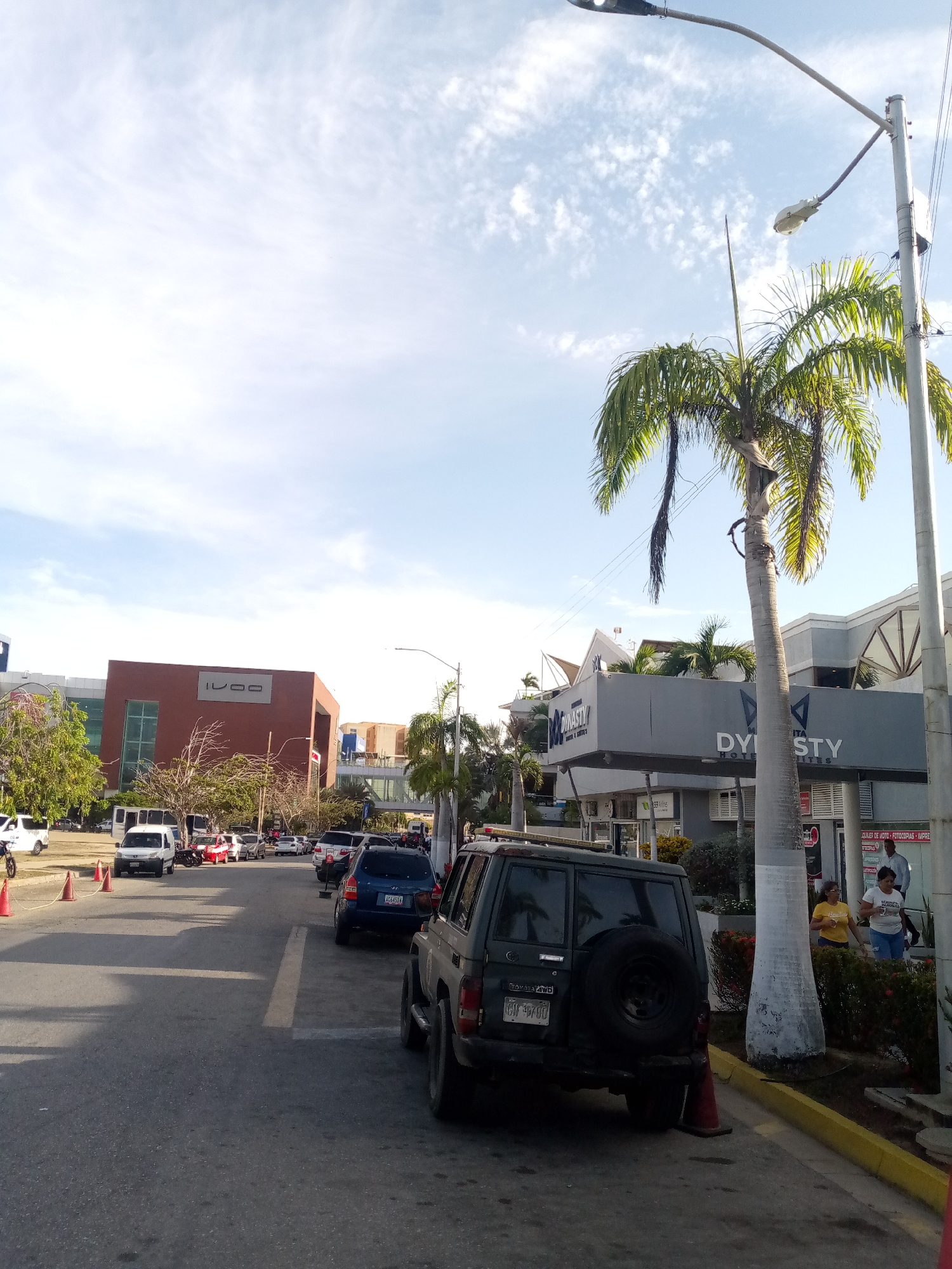 The image captures an urban street scene in Porlamar, Venezuela. The foreground is dominated by a dark green Toyota 4WD vehicle parked on the right side of the street, near the curb. An orange traffic cone is positioned behind the car and partially under the rear bumper. Adjacent to the vehicle is a palm tree, near the Dynasty Hotel & Suites building, identifiable by its signage. Two women are near the base of the palm tree.
The street is lined with several cars parked on both sides, with the traffic flow appearing to move in the direction away from the camera. In the background, on the left side of the street, stands a large, modern building with "IVOO" prominently displayed on its facade. A pedestrian bridge or walkway connects to this building. The sky is partly cloudy, with light blue visible between the scattered clouds. A street light pole, equipped with two lamps, stands tall on the right side.