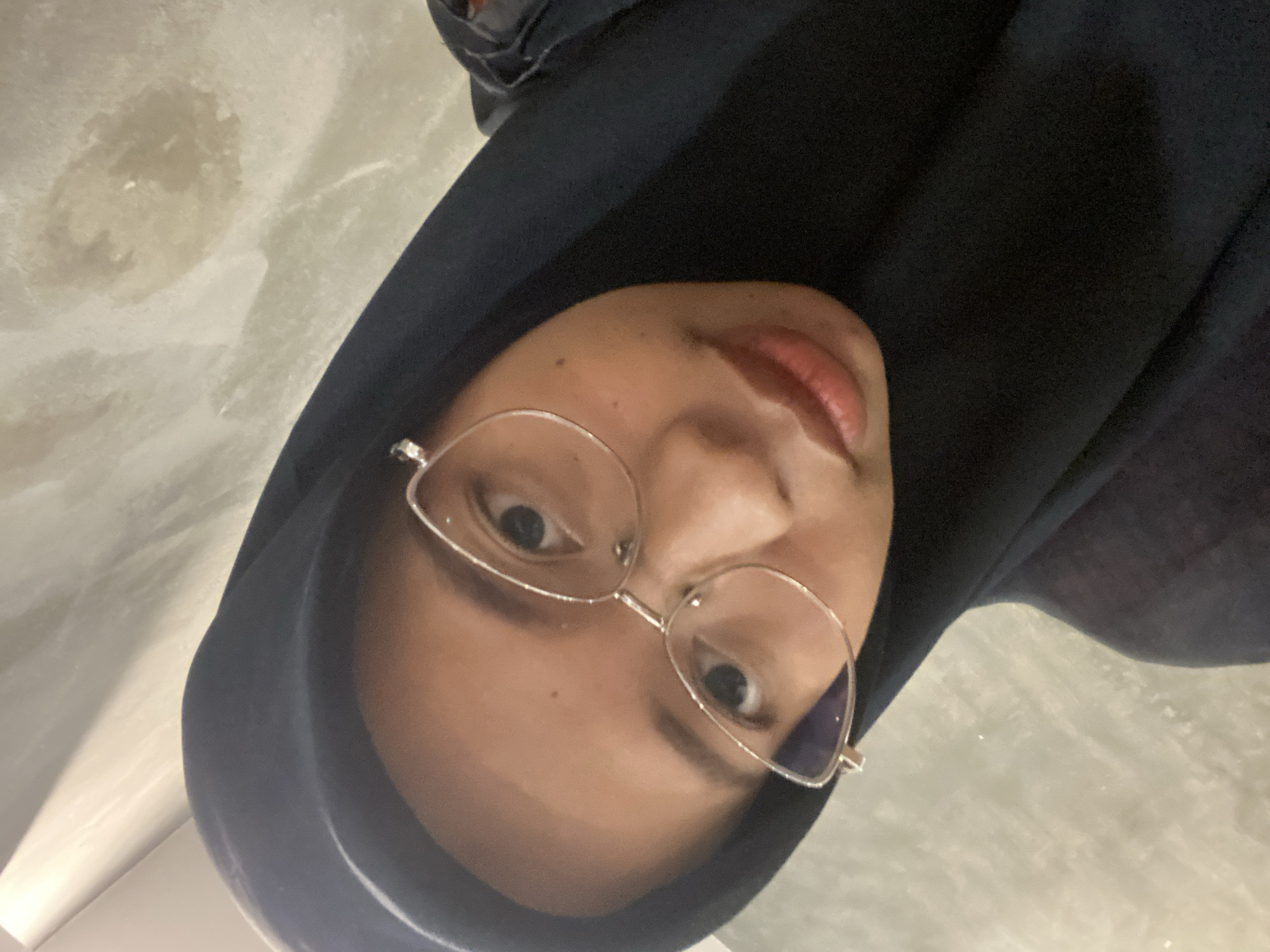 A young woman wearing a black hijab and cat-eye glasses is pictured in a close-up, angled selfie. Her skin tone is light brown, and she has a small mole on her cheek. The background appears to be a textured, light-colored wall, possibly marble or a similar stone. The lighting is dim, casting a slight shadow on her face. The image was taken indoors. No specific location in Sekar Mawar, Indonesia, can be identified from the visual cues in the image.