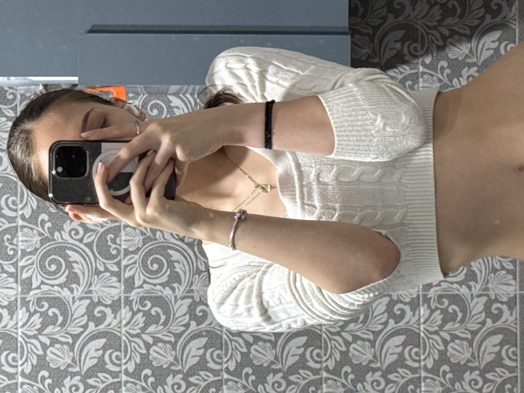 The image, rotated 90 degrees counter-clockwise, captures a person taking a mirror selfie indoors, likely in a bathroom or dressing area located in Sofia, Bulgaria. The individual holds a dark smartphone with both hands, positioned to cover most of their face, with only a portion of their forehead and one eye visible. Their dark hair is neatly pulled back.
The person is dressed in a white, cable-knit, long-sleeved, cropped sweater that reveals their midriff. Accessories include a gold-colored chain necklace, a black beaded bracelet on their upper left wrist (as viewed in the rotated image), and a silver-toned bracelet with a pink charm on their right wrist (as viewed in the rotated image). A ring is also worn on a finger of the hand holding the phone.
The background features a tiled wall adorned with an intricate grey and white floral and scroll pattern. Reflections in the mirror show abstract grey-blue surfaces. The scene is illuminated by indoor lighting, and no specific time of day or weather can be determined. There is no visible text in the image. The person's expression is obscured by the phone.