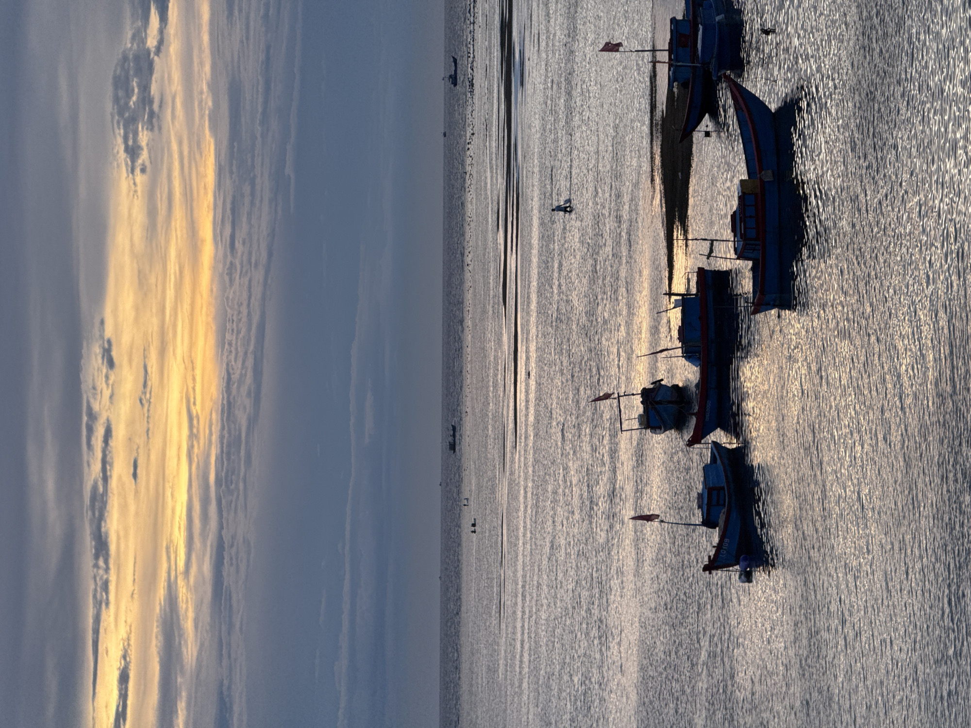 The image shows a serene seascape at dusk, likely taken from the shore of Lý Sơn Island, a Special Administrative Region of Vietnam. The sky is a dramatic canvas of grays and blues, with a band of golden light peeking through the clouds near the horizon, suggesting the sun is setting or has just risen. The water is calm, reflecting the muted colors of the sky, with gentle ripples distorting the light.
In the foreground, several small fishing boats are anchored. They are predominantly blue with red accents, and some have small flags planted on them. The boats are silhouetted against the water, making their details indistinct. The water depth varies, with some boats appearing to be in shallow areas or near sandbars, indicated by darker patches in the water.
Further out in the water, a few individuals are visible. One person is walking in the water, appearing to be a fisherman or local resident. There are also some larger vessels in the distance, possibly larger fishing boats or ships, appearing as dark shapes against the horizon. The overall atmosphere is one of tranquility and quietude, characteristic of a coastal scene at the end of the day. The scene is natural and unadulterated.