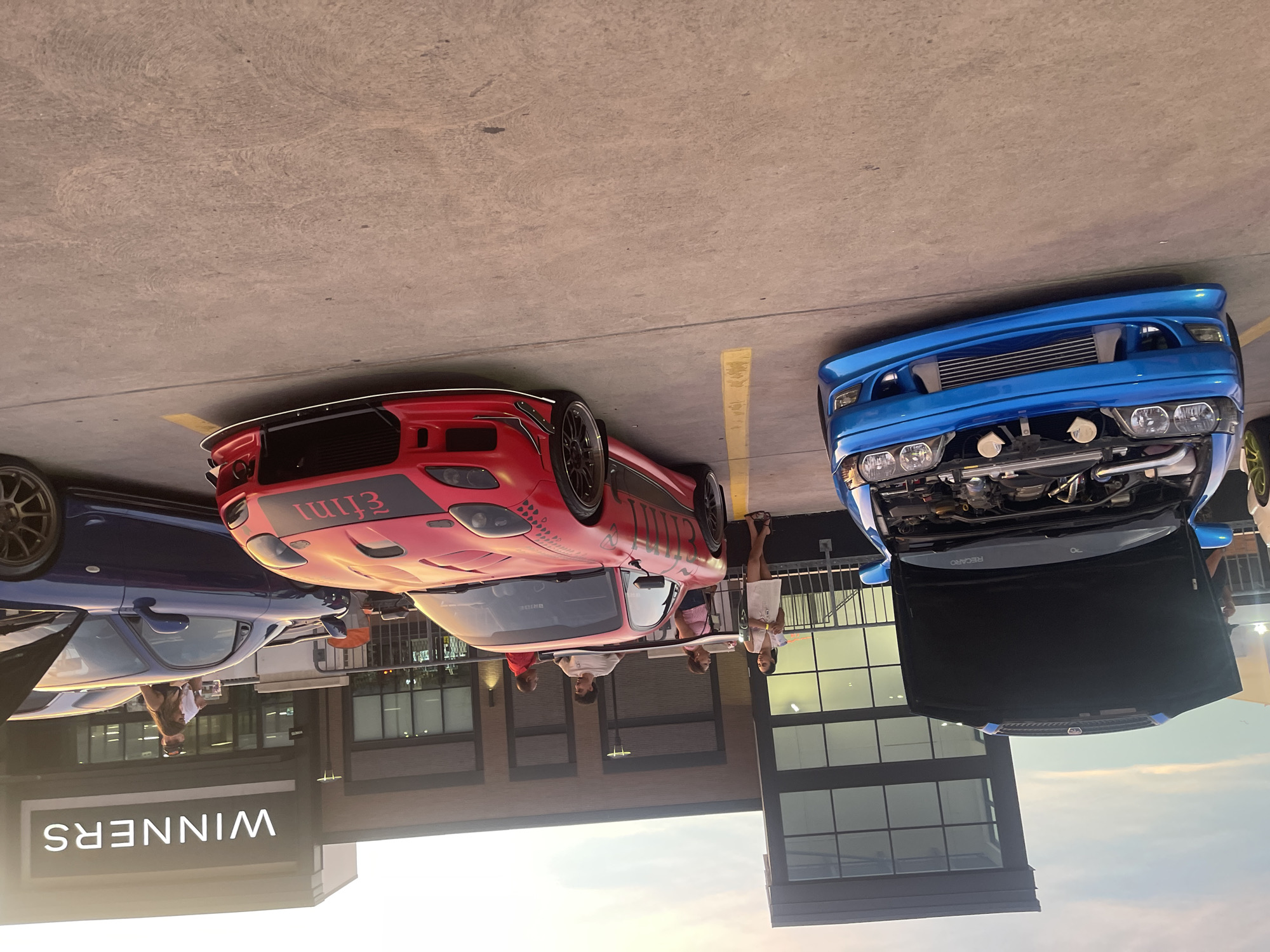 In this outdoor, daylight scene, a car show is taking place in front of a modern building with "WINNERS" signage. The ground is paved concrete, with yellow parking lines visible.
In the foreground, a bright blue car with its hood open is parked on the left. Its engine is exposed, revealing various components. To the right of the blue car is a vibrant red sports car with custom body modifications and graphics. Its front end is visible, and it appears to be a highly customized vehicle. Further to the right, a dark blue sports car is parked, also with its hood open.
Several individuals are present, scattered around the vehicles. On the left, near the blue car, a person is partially visible. In the middle ground, three people are standing and interacting near the red sports car. One individual is holding a drink. Further to the right, two people are standing near the blue sports car, one of whom is holding a drink. The sky is visible in the background, with a light haze suggesting either early morning or late afternoon.
The scene appears to be a casual gathering of car enthusiasts. No specific landmarks definitively place this location in Toronto, Canada, but the architecture and general ambiance are consistent with urban or suburban commercial areas.