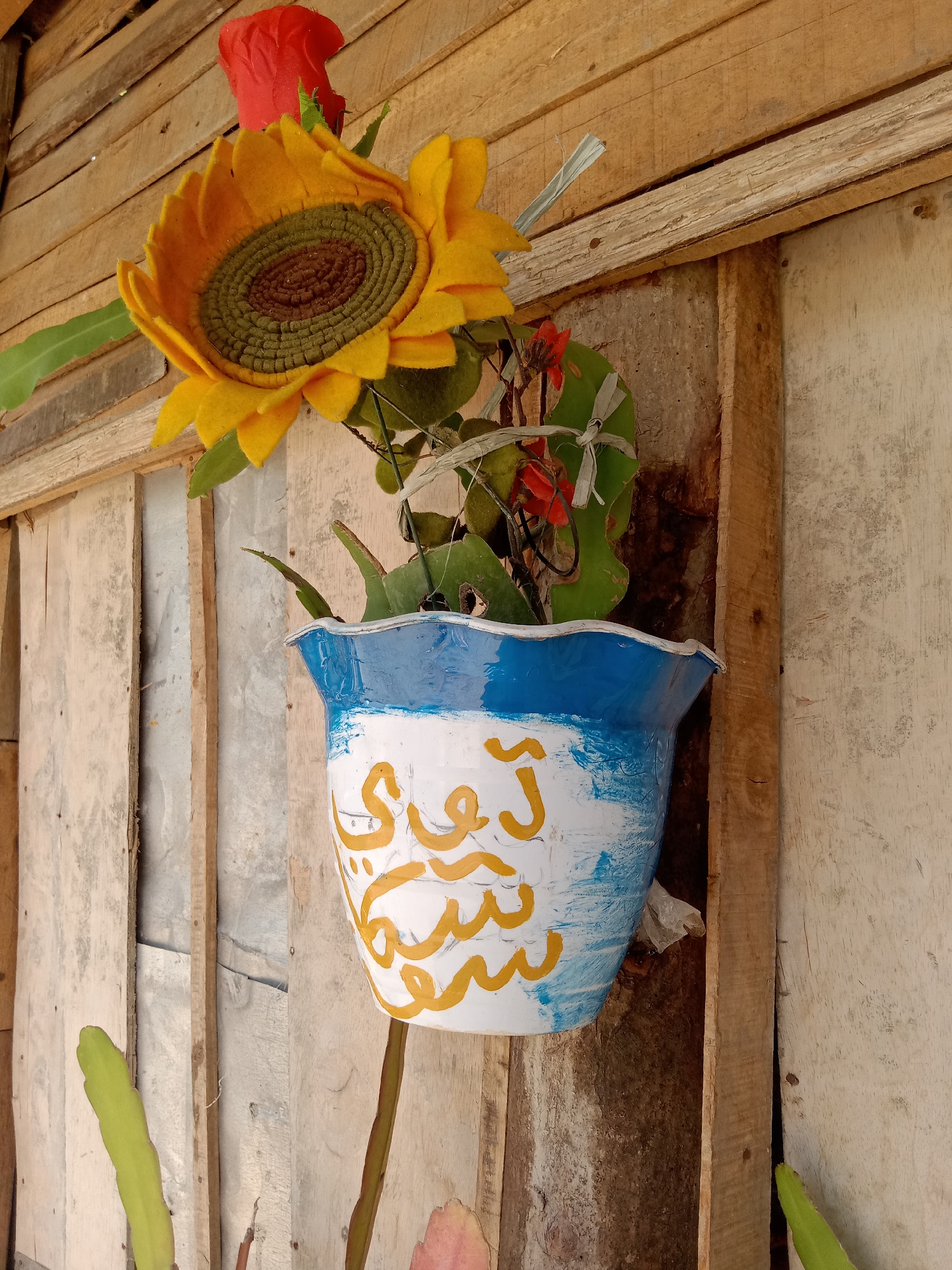 The image shows a close-up of a decorative flower arrangement hanging on a wooden wall. The arrangement consists of a large, artificial sunflower with yellow petals and a brown and green center, and a red artificial rose above it. They are placed in a white pot with blue painted accents and yellow Arabic calligraphy. The pot is attached to a rustic wooden wall made of vertical planks. Some green cactus-like plants are visible at the bottom of the frame, extending from the pot. The lighting suggests it is daytime. The scene is located in Stiaji, Indonesia.