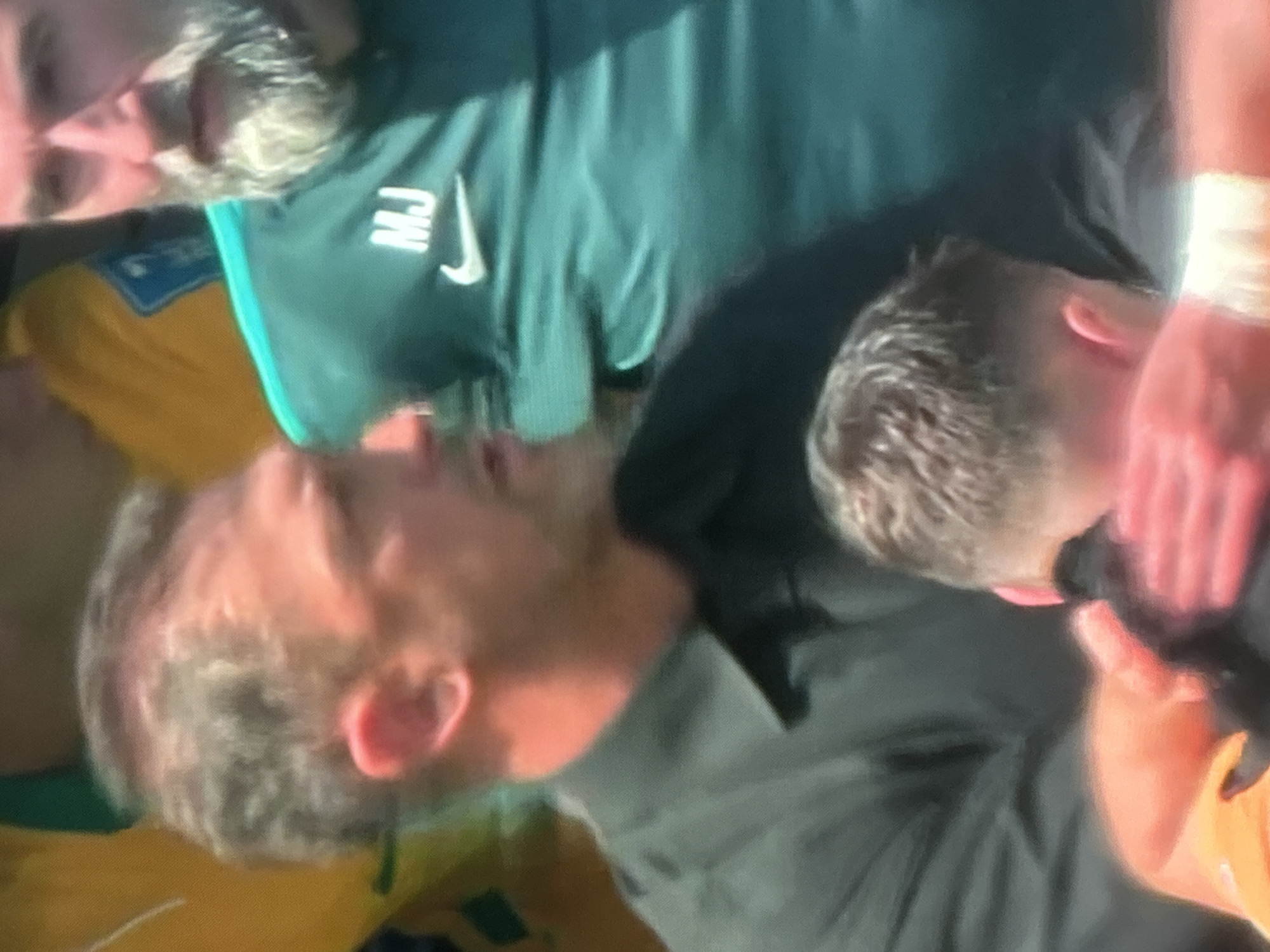 In a close-up, slightly blurry outdoor shot, an adult male with graying, short hair and light skin is seen from the chest up, facing left and slightly away from the camera. He is wearing a black collared shirt. His left arm is visible, and his hand is resting on the head of another person, whose dark hair is partially visible in the foreground.
Behind him, another adult male with a gray beard and light skin is visible from the chest up, facing forward. He is wearing a teal-colored jacket with the white letters "MJ" and a Nike logo on the chest. His facial expression is neutral.
The background is out of focus and appears to be an outdoor setting with blurred figures and colors, possibly a sporting event or gathering. The lighting suggests it is daytime, with warm tones illuminating the skin of the subjects.