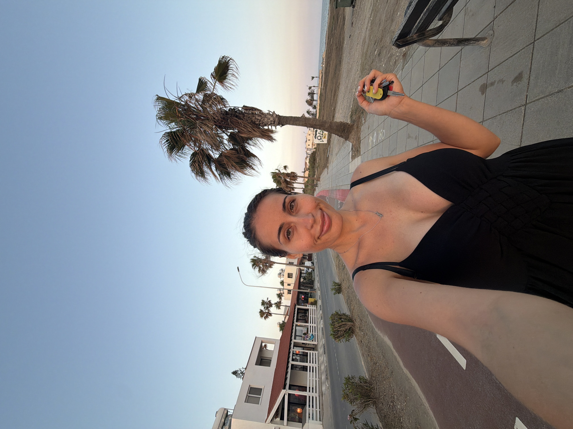 A woman is taking a selfie on a paved walkway next to a road. She is wearing a black dress and a necklace. She is holding a set of keys in her right hand. In the background, there are buildings with white walls and red roofs, palm trees, and the sea. The image appears to have been taken in Meneou, Cyprus, given the architectural style and vegetation. The lighting suggests it is either early morning or late afternoon.
