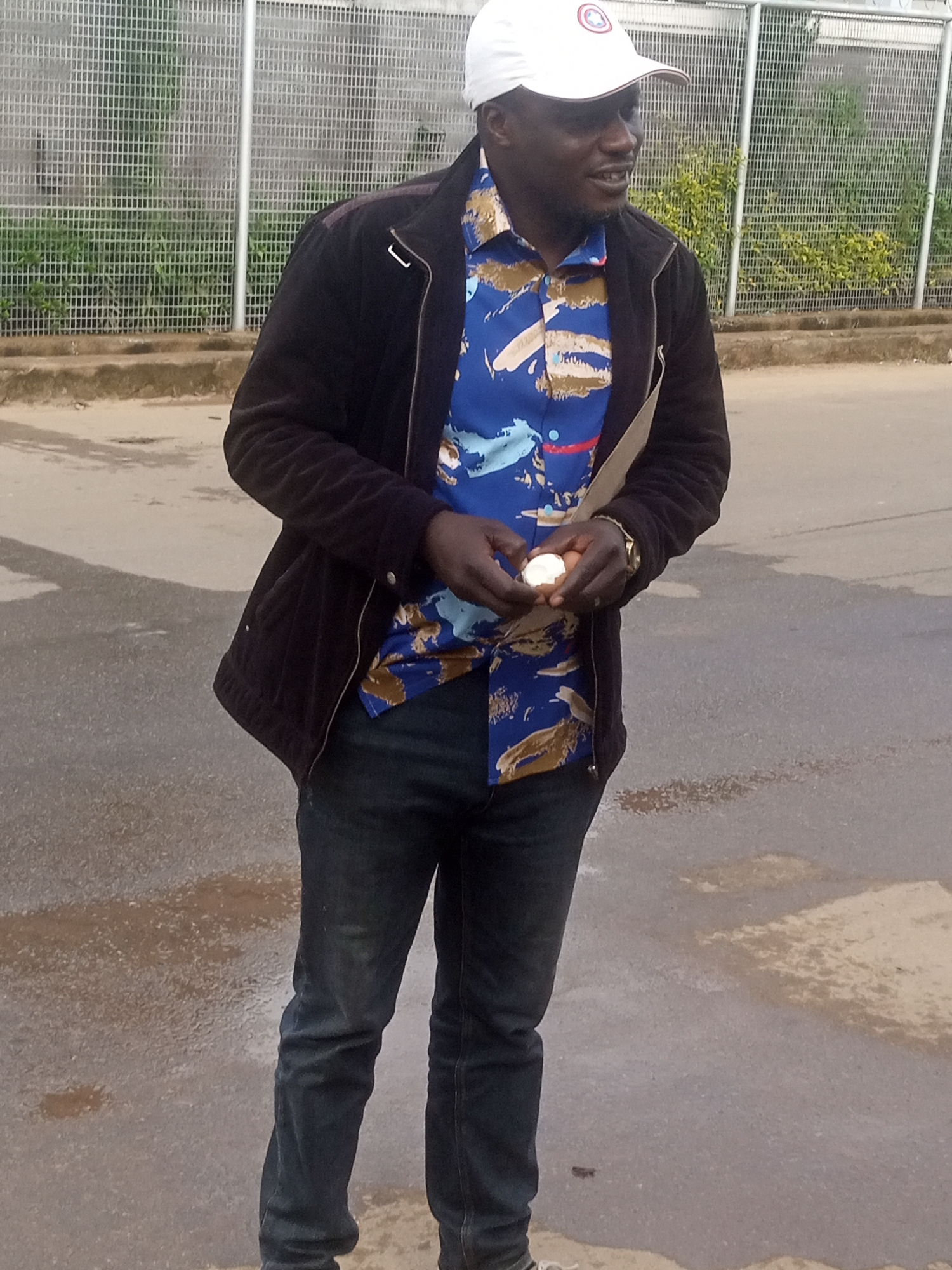A medium shot captures a Black man standing outdoors on a wet asphalt road in front of a metal fence. He wears a white baseball cap, a dark jacket over a blue and brown patterned shirt, and dark blue jeans. His right hand holds a white egg. The man appears to be speaking, with his mouth slightly open. The background shows a grassy area behind the fence. The lighting suggests it might be overcast or a cloudy day.