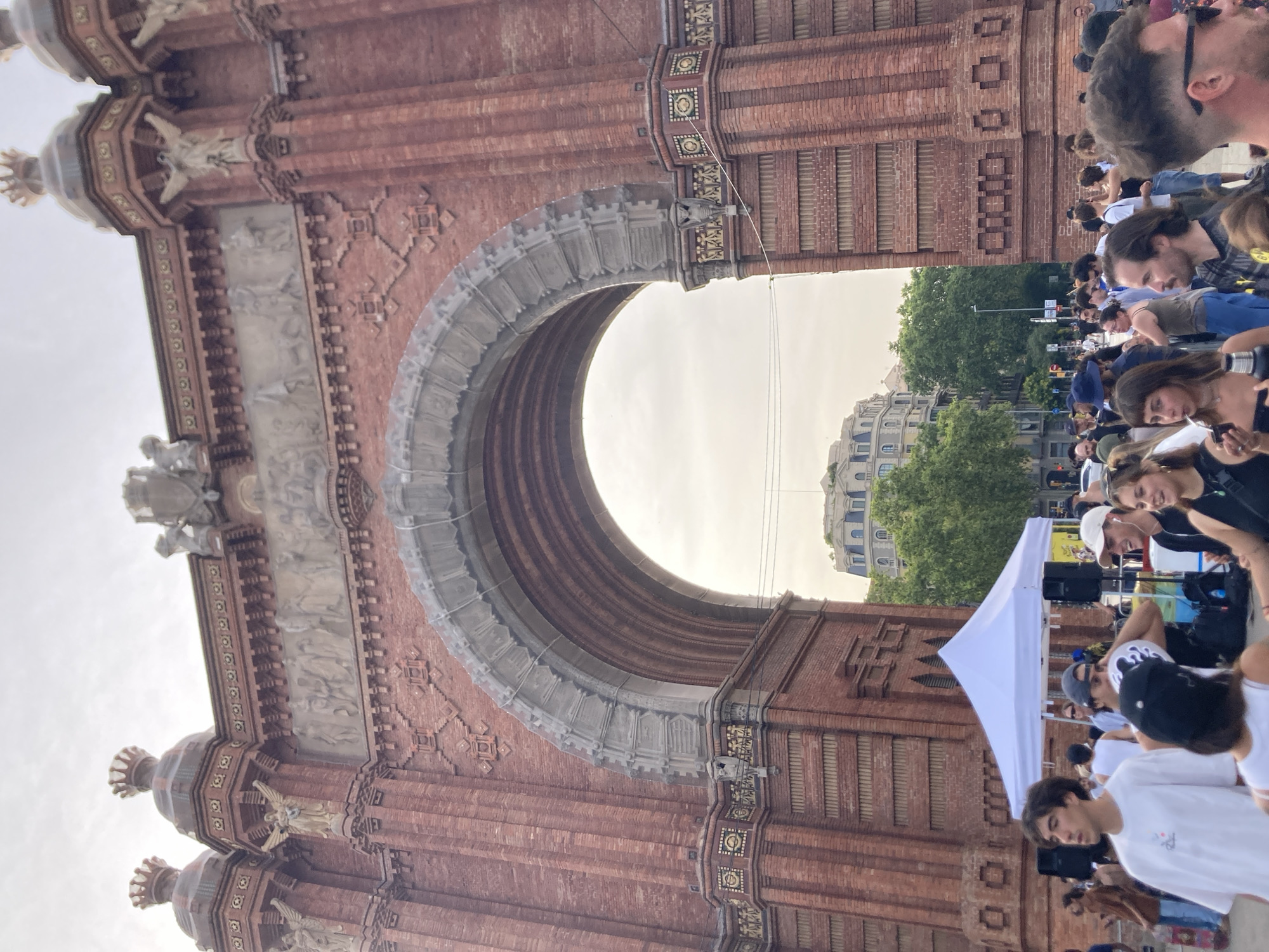 The image depicts the Arc de Triomf in Barcelona, Spain. The monument is constructed of red brick and features intricate carvings and sculptures. In the foreground, a crowd of people is gathered, some standing near a white pop-up tent. Several individuals are looking in different directions, suggesting an event or gathering is taking place. The sky is overcast. In the background, beyond the arch, a building with a domed roof and trees can be seen. The overall scene suggests a public space with an active presence of people.