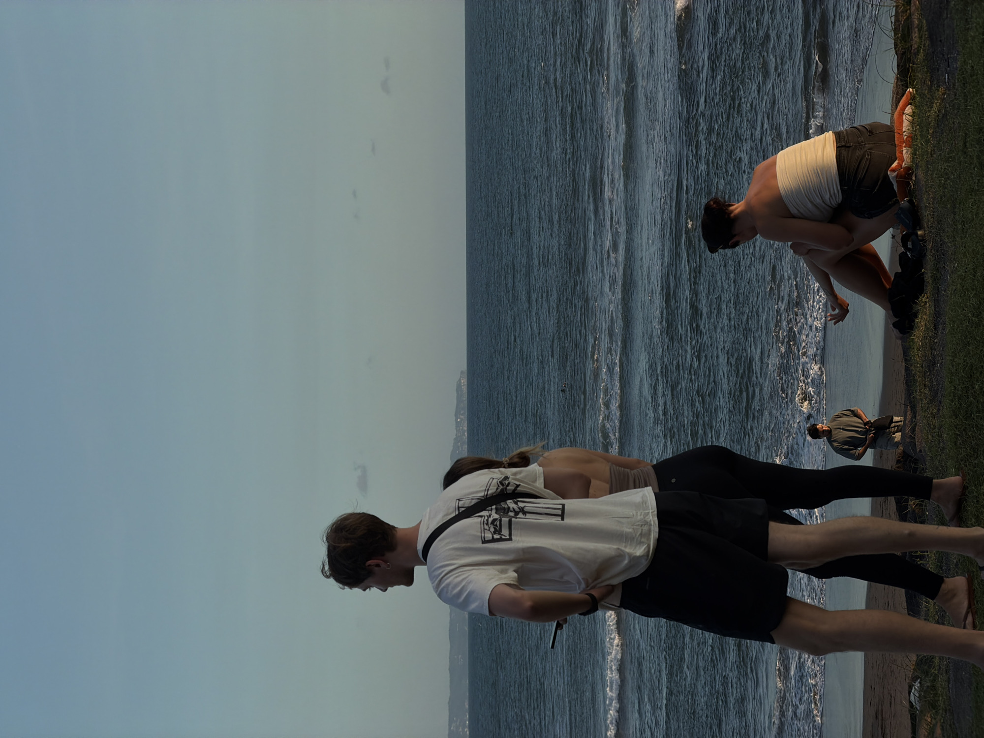 The image shows a coastline, possibly in Mengwi, Indonesia, given the presence of a coastline and local residents. In the foreground, two people are standing near the water's edge. One individual wears a white t-shirt with a graphic design and black shorts. The other individual wears a tan top and black leggings. Further towards the right edge of the image, there is another person bending over near the water, wearing a white top and dark shorts, with black footwear. On the right side, at the edge of the grass, an orange plush toy is visible. To the right of the plush toy is a man in a green shirt and grey pants, kneeling. The background is filled with a body of water, potentially the ocean, and a clear sky above.