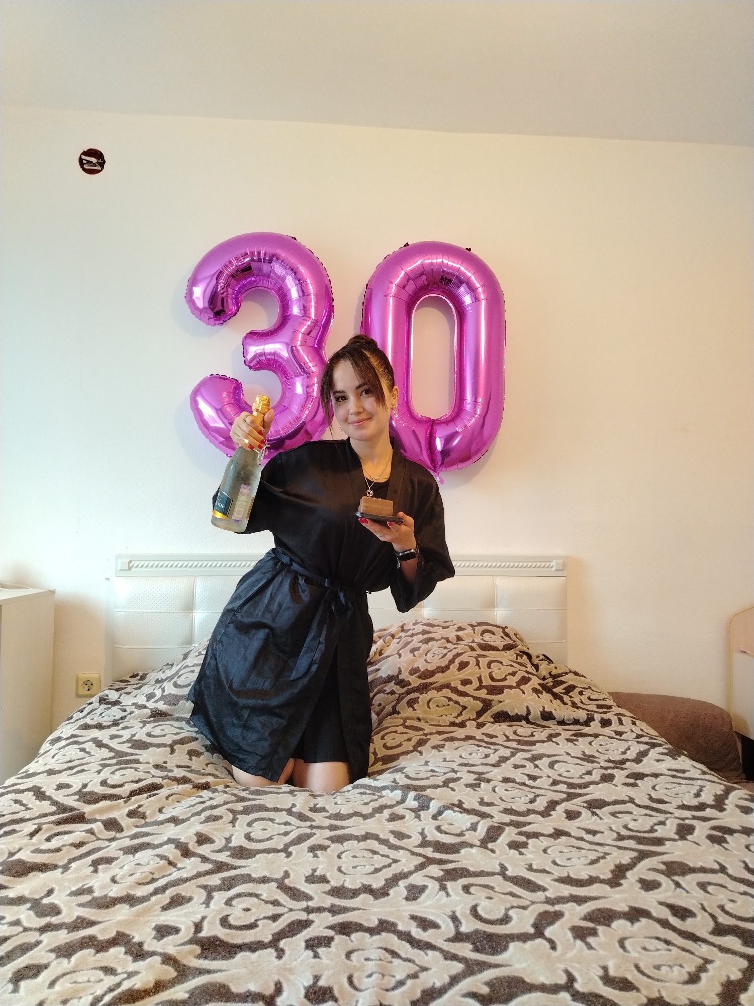 A woman kneels on a bed in a room, holding a bottle of champagne and a slice of cake. Behind her, two large, pink balloons shaped like the numbers 3 and 0 are attached to a white wall. The bed is covered with a patterned comforter. The woman is wearing a black robe. A light switch and an electrical outlet are visible on the wall. There is a bedside table on the left and a headboard behind the woman. The room appears to be a bedroom. The location is not specified in Noviy, Russia.