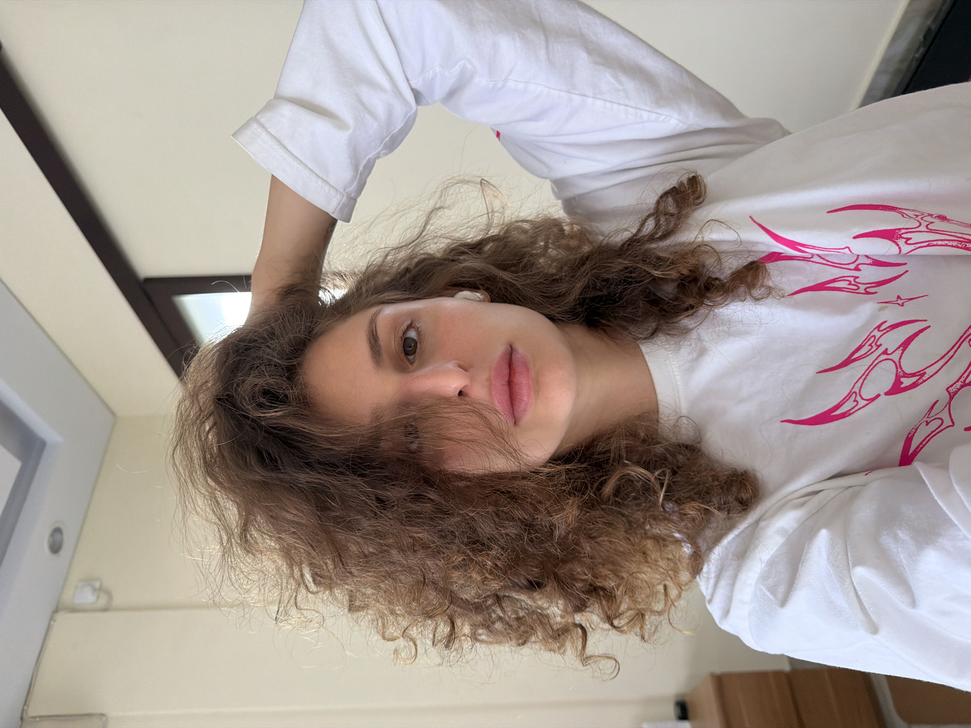 A selfie-style portrait depicts a young adult female, likely in her late teens or twenties, positioned indoors. The subject has voluminous, light brown, curly hair, some strands of which partially obscure her right eye. She has a neutral expression, with full, slightly parted lips and a visible left eye gazing directly into the camera. A white wireless earbud is present in her left ear. She wears a white long-sleeved t-shirt featuring a prominent bright pink graphic print on the chest, characterized by stylized flame-like or abstract shapes. Her left arm is raised, with her hand positioned at the back of her head, appearing to touch or adjust her hair.
The background consists of light-colored, possibly off-white or beige walls. In the upper left quadrant, a white rectangular ceiling light fixture and a circular recessed light are visible, along with a small white wall-mounted device and a thin wire. To the right, a portion of a dark-framed opening, possibly a window or doorway, is visible, offering a glimpse of bright light. A dark horizontal beam runs above this opening. In the lower-left corner, a segment of a light brown wooden furniture piece, consistent with a dresser or bedside table, is partially visible. The lighting is bright and even, suggesting a well-lit indoor environment. The location is identified as North Kuta, Indonesia, though the image itself does not contain specific visual indicators to confirm this.