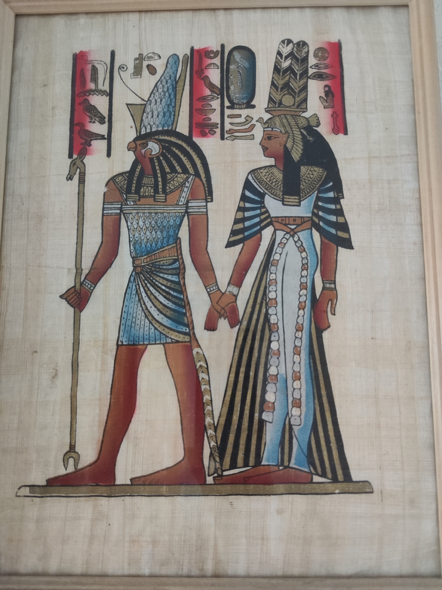 This image is a framed papyrus painting depicting two figures in ancient Egyptian style. The figure on the left is a male with the head of a falcon, wearing a blue and white patterned kilt, a broad collar, and holding a staff. The figure on the right is a female, with darker skin and elaborate headdress, wearing a long white dress with patterned sleeves and a decorative fringe. They are holding hands. The background features hieroglyphs and decorative elements. The painting is framed with a light-colored wooden frame and is presented as an artifact. There is no discernible location or context within the city of Astana, Kazakhstan, as the image purely depicts an artistic representation of ancient Egyptian culture.
