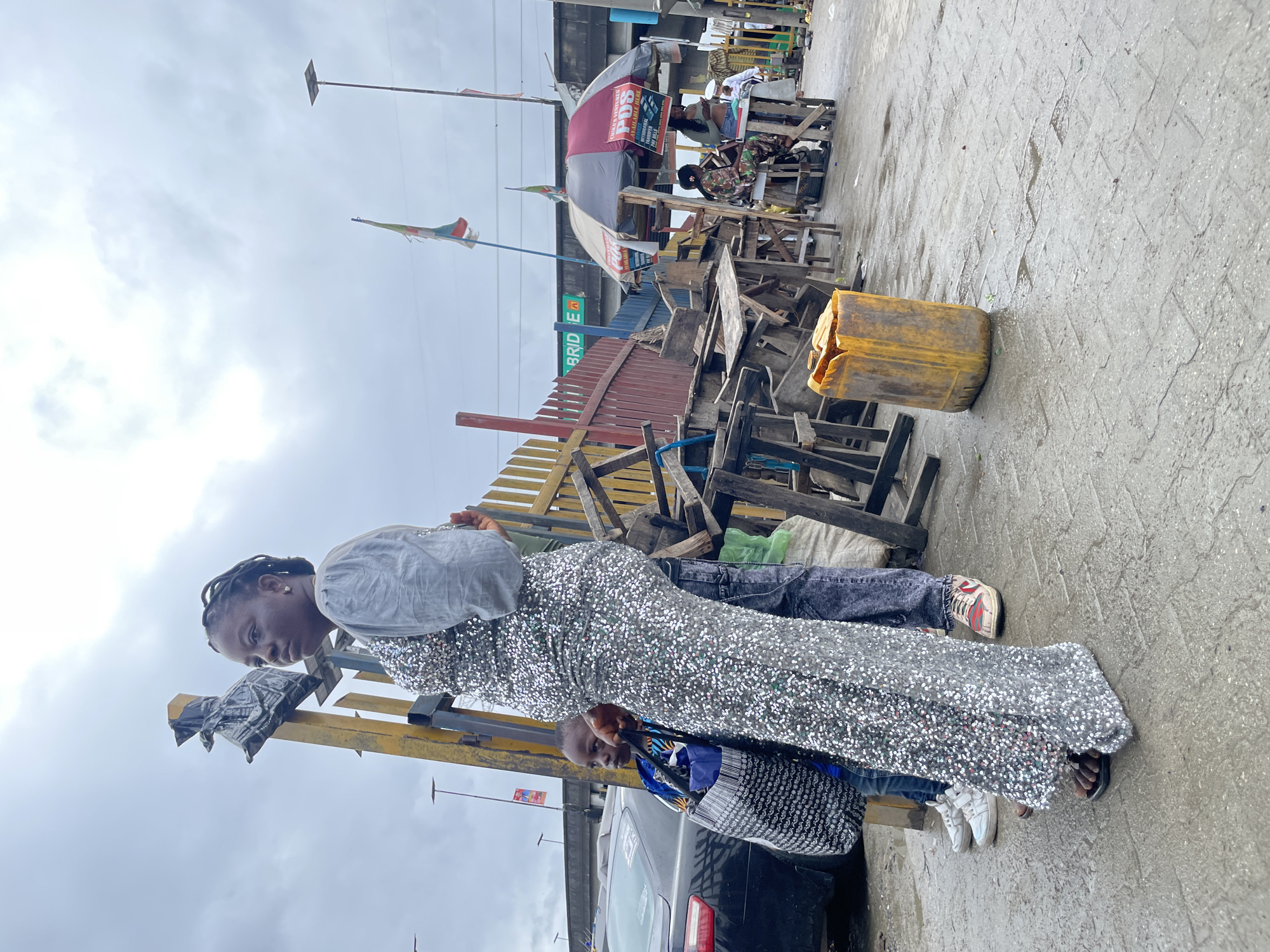 In Ajah, Nigeria, a woman in a shimmering silver dress and a young boy stand near a yellow jerrycan and stacks of wooden tables and chairs. The ground is wet, indicating recent rain. In the background, a black car is parked, and various market stalls are set up under umbrellas. Flags are visible, and an overhead bridge with a sign reading "BRIDGE A" is present. The sky is overcast.