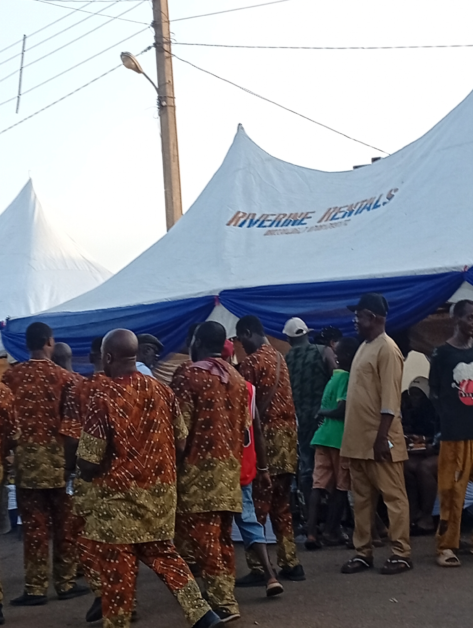 A group of men are gathered outdoors in what appears to be a public event or festival in Nsugpe, Nigeria. They are standing in front of a large white tent with blue draping, and the tent displays the text "RIVERINE RENTALS". Several men are wearing matching traditional brown and gold patterned clothing, suggesting a cultural or celebratory gathering. Some are facing away from the camera, while others are looking around. The environment includes a utility pole with wires overhead and a bright, clear sky. The ground is paved with what looks like asphalt or concrete.