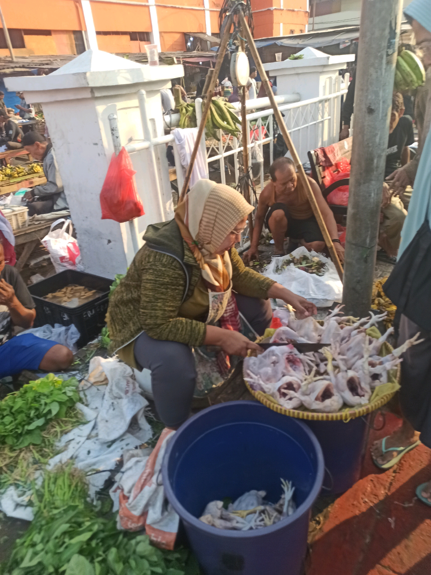The image captures a bustling scene at an outdoor market, likely in Cikarang, Indonesia, with a focus on a woman vendor in the foreground. She is wearing a headscarf and an olive-green jacket over a lighter green garment, and is engaged in preparing poultry. In front of her, a woven basket is filled with raw chickens, and she is holding a knife, seemingly in the process of cutting or cleaning them. To her right, a large blue plastic tub contains more chickens. To her left, a black plastic container holds what appears to be fish. Behind her, a man is crouched near a pile of what seems to be dried leaves or fodder, and further back, a red plastic bag hangs from a wooden structure. The environment is characterized by a mix of market stalls, concrete structures, and vegetation, suggesting a busy commercial area. Other individuals are visible in the background, engaged in their respective activities, contributing to the lively atmosphere of the market. The lighting suggests it is daytime, possibly morning or late afternoon, with warm hues casting a soft glow on the scene.