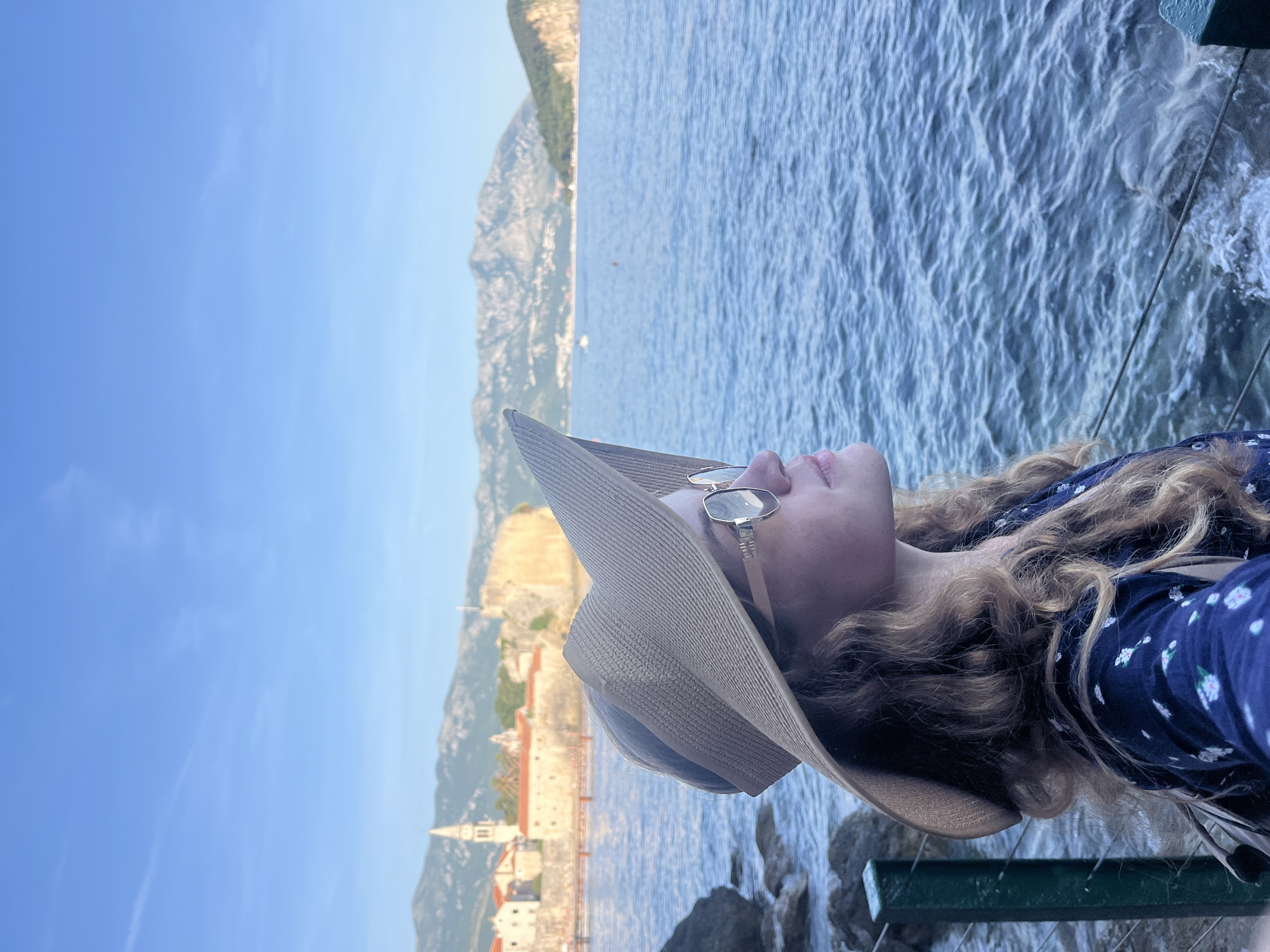 A picturesque coastal scene captures a young woman in profile, looking upwards, on what appears to be a sunny day. The image is framed with the woman occupying the right side, showcasing her long, curly blonde hair, a wide-brimmed straw hat, and stylish sunglasses. She is wearing a dark blue top adorned with a white floral pattern.
To her left, the calm, blue sea stretches across the mid-ground, displaying subtle ripples on its surface. Beyond the water, in the distance, lies a historic old town or fortress, characterized by its ancient stone walls, traditional red-tiled roofs, and a distinctive bell tower. Towering mountains provide a majestic backdrop to the town, rising under a clear, bright blue sky with sparse, faint clouds.
In the immediate foreground, a dark green railing or fence and some rocks indicate the woman is standing close to the water's edge, possibly on a promenade or viewing platform. The overall atmosphere is one of peaceful enjoyment and appreciation for the beautiful scenery. The specific architectural cues of the old town and the dramatic coastal landscape, coupled with the provided context, confirm the location as Budva, Montenegro. There is no visible text in the image.