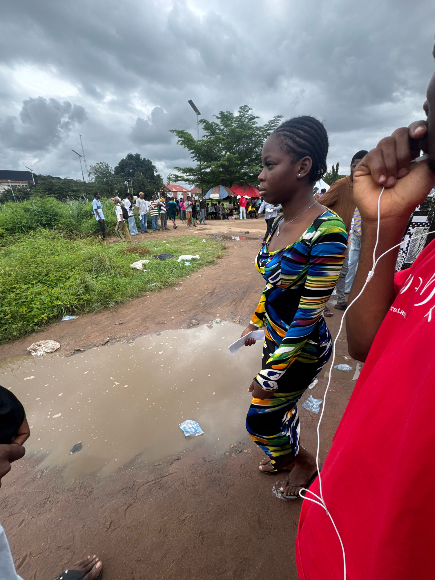 In Ihiagwa, Nigeria, a woman stands in profile on a dirt road that is partially flooded with muddy water, reflecting a cloudy sky. She is wearing a long-sleeved dress with a vibrant, abstract pattern of blues, greens, yellows, and black. Her hair is styled in braids. In her right hand, she holds a white piece of paper. She is looking to her left.
Behind her, a crowd of people are gathered. Some are standing, while others are seated under umbrellas. The area appears to be an outdoor market or gathering space. The ground is uneven, with patches of grass and debris visible. In the foreground, a person wearing a red shirt is partially visible on the right, with white earbuds and a wire extending from their ear. The overall scene suggests a bustling, possibly informal, urban environment.