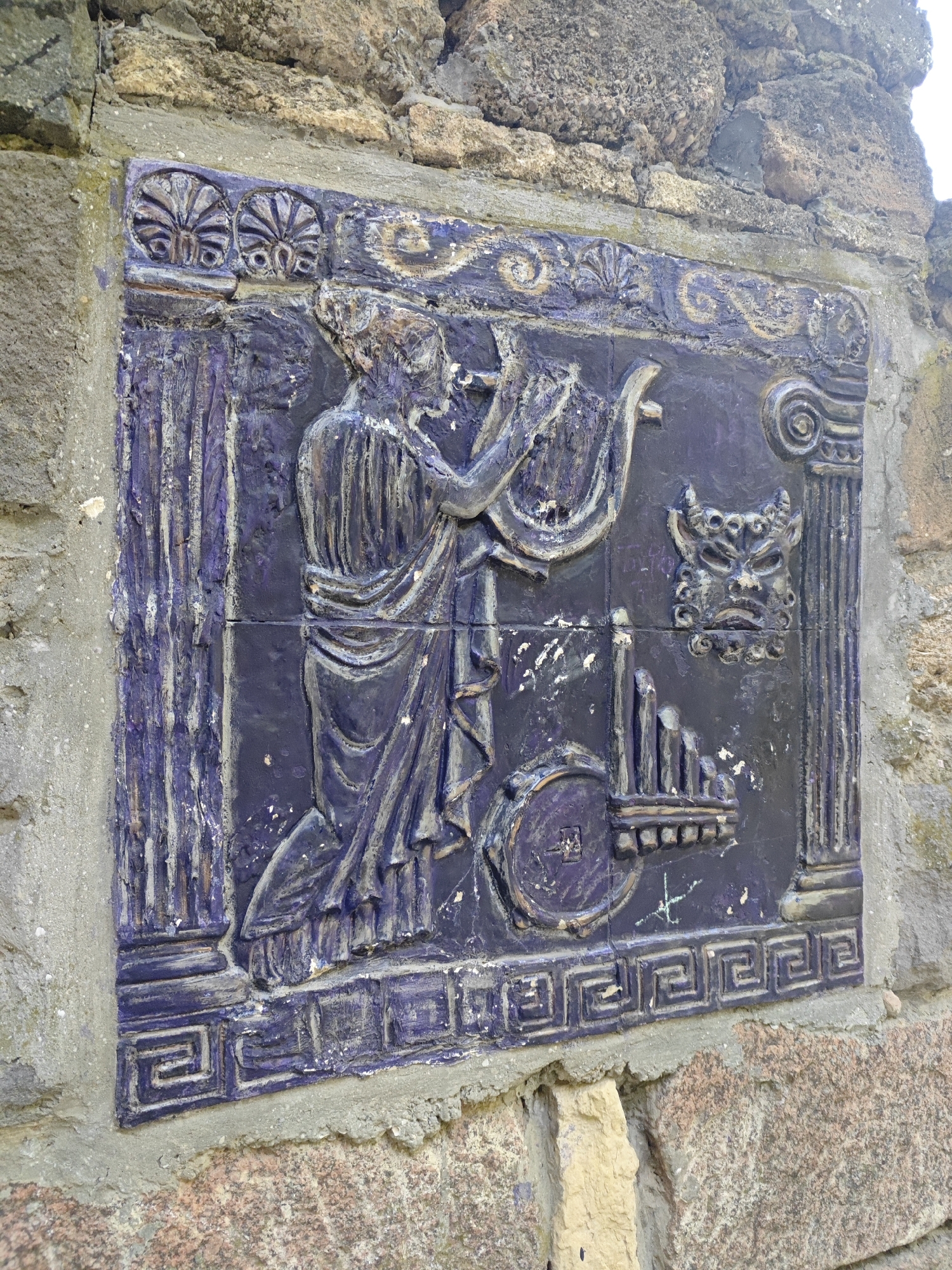 This is a medium shot, eye-level, outdoor photograph of a dark blue, relief-carved ceramic tile affixed to a stone wall. The tile is ornately decorated with figures and symbols that appear to be of ancient Greek or Roman origin. The central figure, depicted in the upper left quadrant of the tile, is a person in flowing robes, kneeling and playing a lyre. To the right of this figure, there is a grotesque mask with horns and a menacing expression, followed by a column with a volute capital. Below the mask and column is what appears to be a depiction of aulos pipes, a double-reed instrument. The bottom border of the tile features a Greek key (meander) pattern. The tile is slightly worn and chipped in places, with white paint or plaster residue scattered across its surface. The surrounding wall is constructed of rough-hewn stones of varying sizes and colors, ranging from tan and beige to reddish-brown, with visible mortar. The upper portion of the wall is rougher and more irregular in texture. The lighting suggests it is daytime, with some shadows indicating the sun's position. The scene is static and appears to be a historical or artistic installation.