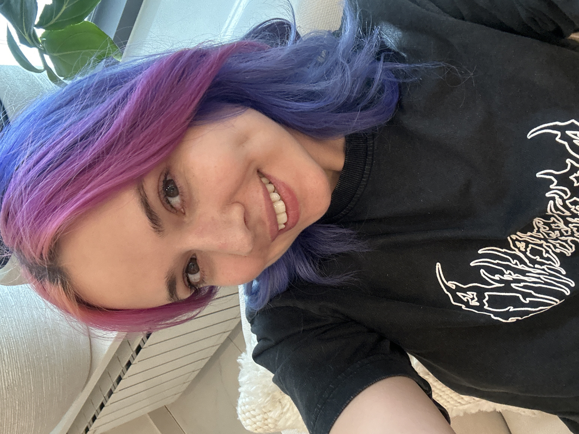 A woman with striking, split-dyed hair, featuring shades of purple and pink on one side and blue on the other, is depicted in a close-up, upward-angled selfie. She is smiling broadly, revealing her teeth, and looks directly towards the camera with warm brown eyes. She wears a black long-sleeved shirt adorned with a prominent white outline graphic on the right side, which appears to be a stylized mythical creature or beast.
The setting is indoors, likely a casual home environment, with a light-colored, textured cushion or blanket partially visible behind her. A green houseplant is also seen in the upper left background, suggesting natural light is present, indicating it is daytime. The woman's expression conveys happiness and cheerfulness. This scene is set in the City of Zagreb, Croatia. No legible text is visible within the image, beyond the intricate graphic design on her shirt.