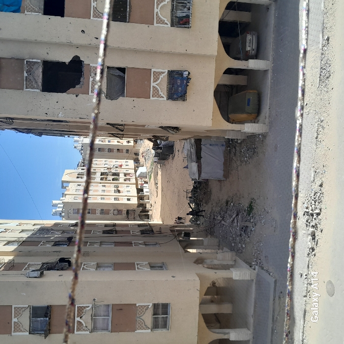 Between two rows of buildings stands a dusty corridor. The structures are beige, multi-storied, and show damage with gaping holes in some windows. Discarded items and debris clutter the ground between them. The blue sky peeks above the buildings on the left side. A road runs perpendicular to the building rows, visible on the right, with a sidewalk littered with debris. The image is rotated clockwise.