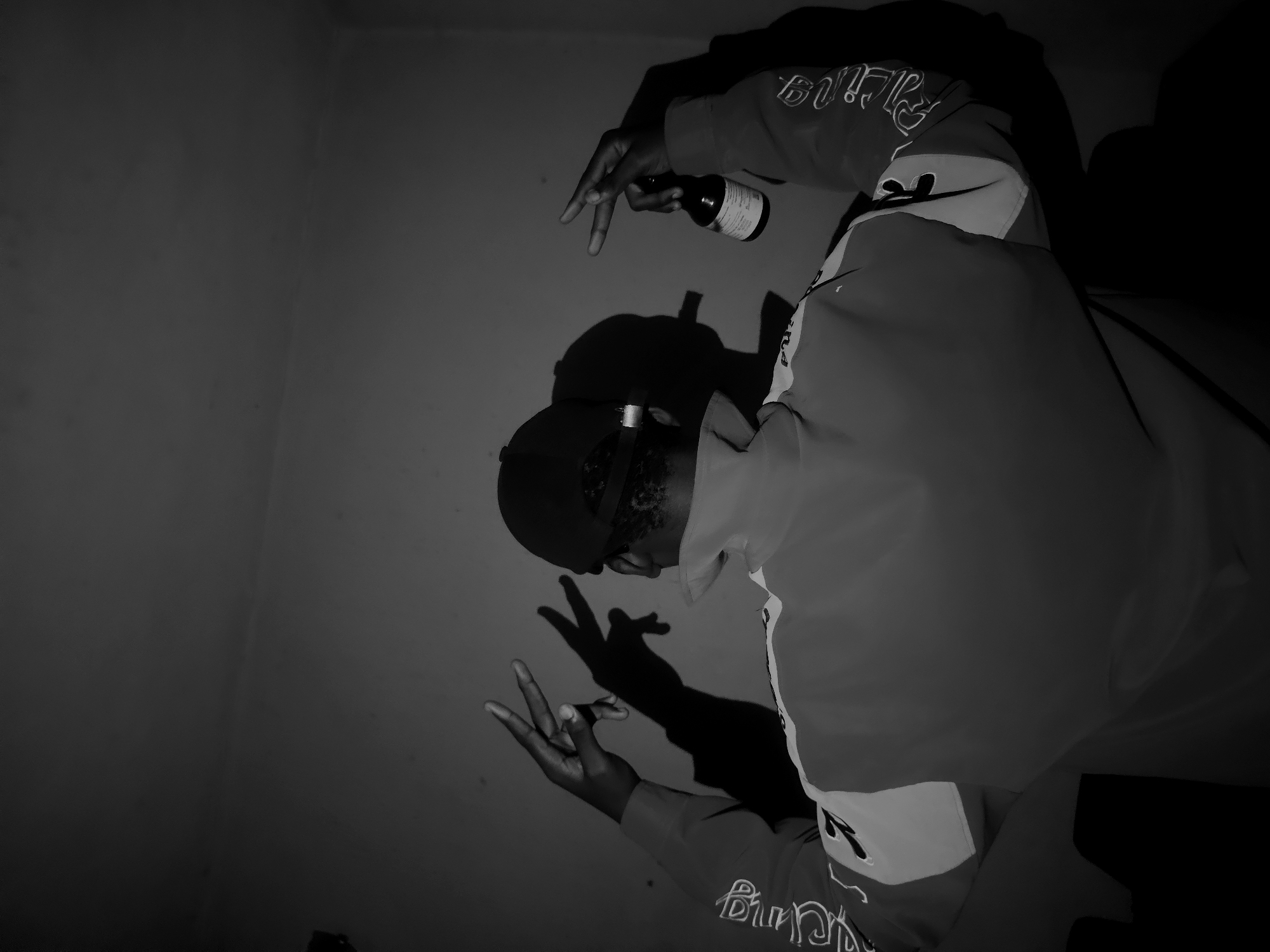 A monochromatic, high-contrast photograph captures a person indoors, likely in Asaba, Nigeria, against a plain light-colored wall. The image is presented with a significant counter-clockwise rotation from its probable original orientation. The subject, viewed from what appears to be a high angle looking down, is wearing a dark cap and a jacket featuring distinct sections and stylized white text on the sleeves.
In their raised right hand, the person holds a small, dark bottle with an illegible label. Their left hand is positioned lower, making a "V" or peace sign gesture. Strong, directional lighting casts sharp, exaggerated shadows of the person and their hands onto the wall, emphasizing their profile, the contours of their hands, and the bottle. The subject's face is obscured by the cap and the camera angle, adding an element of mystery.
Visible text on the jacket sleeves includes stylized white script; on the upper right sleeve, the text resembles "Racing" or a similar word, while text on the left forearm is also in a decorative script. The simple background and dramatic lighting focus attention entirely on the individual and their deliberate pose.
