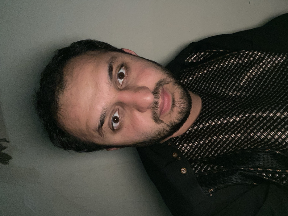 A man with dark hair and a beard is wearing a black and silver patterned shirt with gold buttons. He is looking directly at the camera with a neutral expression. His skin has a slightly ruddy complexion. The background appears to be a plain, light-colored wall, slightly out of focus. The image is oriented with the subject at an angle.