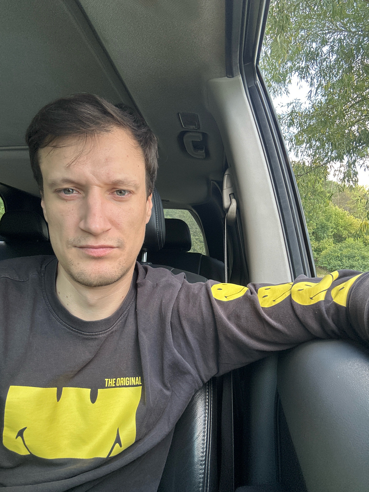 A man with dark hair and light-colored eyes is seated in the driver's seat of a car, looking directly at the camera. He is wearing a dark long-sleeved shirt with a large yellow smiley face graphic on the front, and the text "THE ORIGINAL" above it. The sleeve of the shirt also features a pattern of yellow smiley faces. The car's interior is visible, including the dashboard, headliner, and a seatbelt. Through the open car window, lush green trees and foliage are visible under a bright sky, suggesting an outdoor, possibly rural or park-like setting. The image was taken in Makiv, Ukraine.