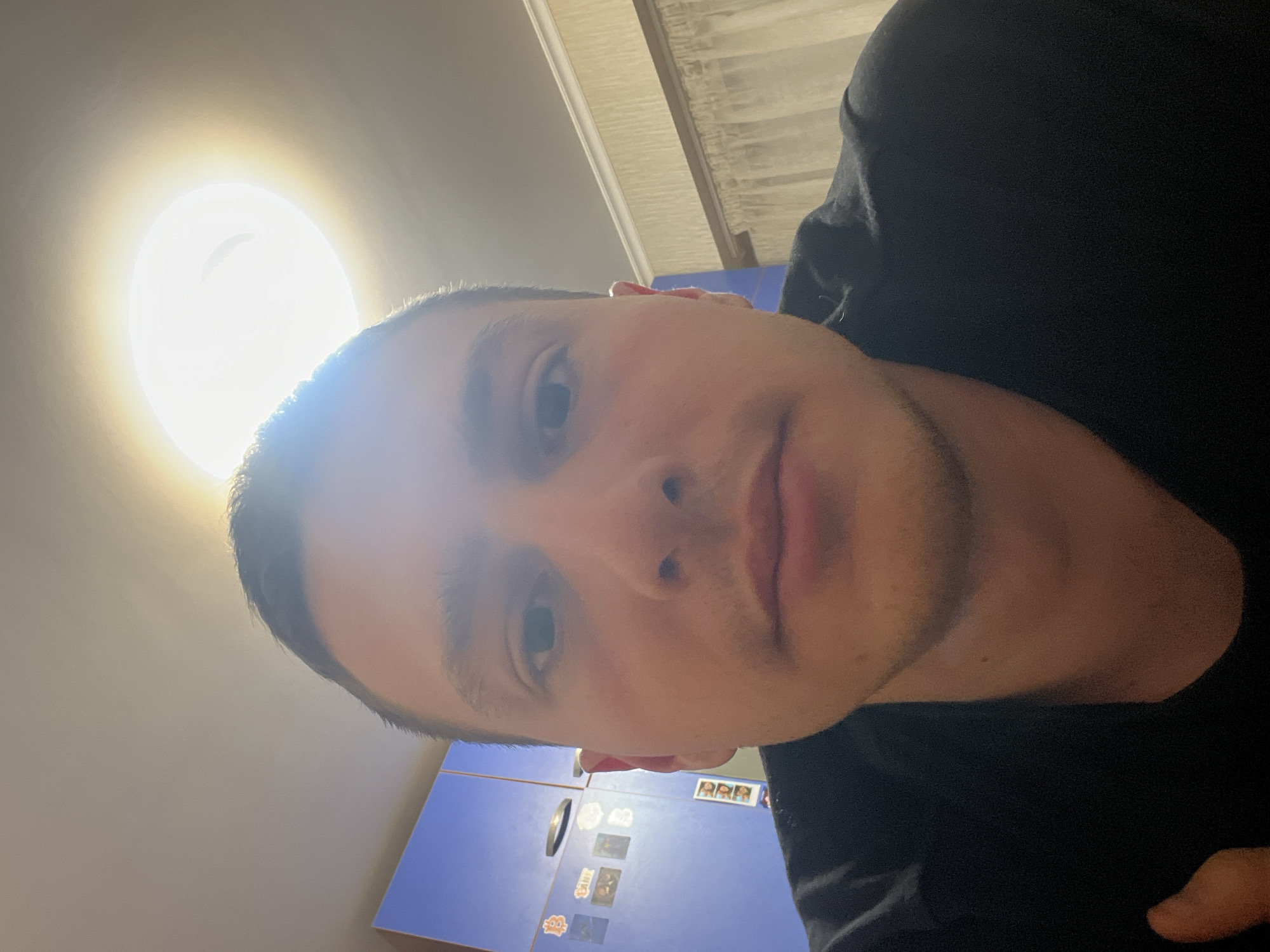 A close-up selfie of a light-skinned male with short brown hair, visible facial stubble, and brown eyes is displayed. He is wearing a black T-shirt. The background includes a bright ceiling light and a blue cabinet adorned with magnets or stickers. Given the location mentioned, it can be assumed this is taken indoors, likely in Burgas, Bulgaria, although specific geographic features are absent.