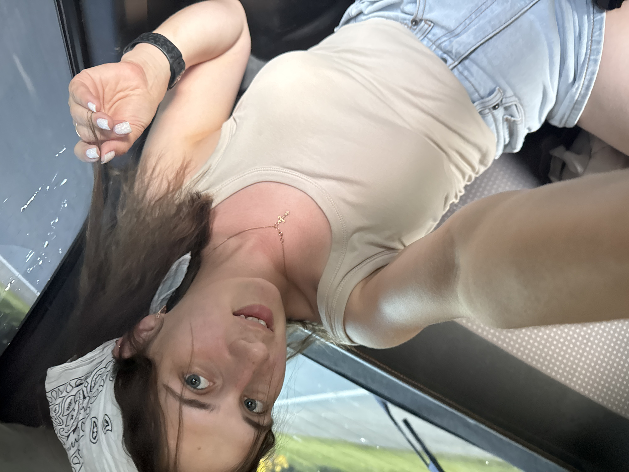 In the image, a young Caucasian woman with brown hair and blue eyes is taking a selfie. She is wearing a beige tank top, light blue denim shorts, a white bandana with black paisley print on her head, and a gold cross necklace. Her fingernails are painted white. She is wearing a black watch on her left wrist. The woman is sitting in what appears to be the back seat of a vehicle, possibly a bus or van, as indicated by the seating upholstery and the view through the window which shows a road and greenery. The overall setting is casual and appears to be outdoors. The specific location in Bol'shaya Sosnova, Russia, cannot be determined from the visual information alone.