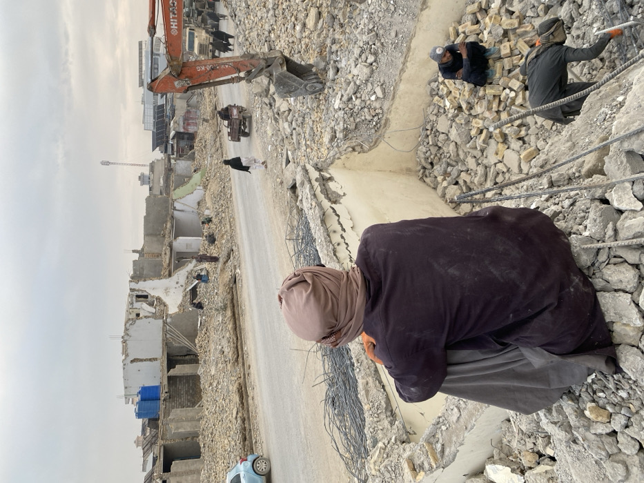 The image captures a scene of extensive urban demolition and debris in Kandahar, Afghanistan, under an overcast sky.
In the immediate foreground, an individual is seated on a large pile of concrete rubble, facing away from the camera. This person wears a dark, long-sleeved garment, dark trousers, and a light brown head covering that conceals the head and neck. Orange gloves are visible on their hands. To the lower right of this individual, two other people are present amidst piles of broken concrete and bricks. One person crouches, wearing dark clothing and a grey beanie, while another, also in dark attire and a light head covering, bends over, appearing to engage with metal rebar and debris.
A paved road runs horizontally across the midground. A light blue compact car is visible on the left side of the road. On the road itself, a person in dark clothing walks away from the camera. Further right, a small, three-wheeled cargo vehicle travels in the same direction. Several other pedestrians in dark clothing are visible near the right edge of the road. A large, orange Hitachi excavator is prominently positioned on the right, surrounded by extensive rubble, its bucket lowered towards the ground.
The background consists of a landscape of heavily damaged and partially demolished multi-story buildings, lining what appears to be a street parallel to the foreground road. These structures exhibit exposed rebar, broken concrete, and fragmented brick walls. Two blue plastic water tanks are situated on the rooftop remnant of a building on the left. In the distant background, a tall communication tower stands, alongside other structures, one of which has visible solar panels on its roof. The entire environment is dominated by demolition debris and the skeletal remains of buildings.