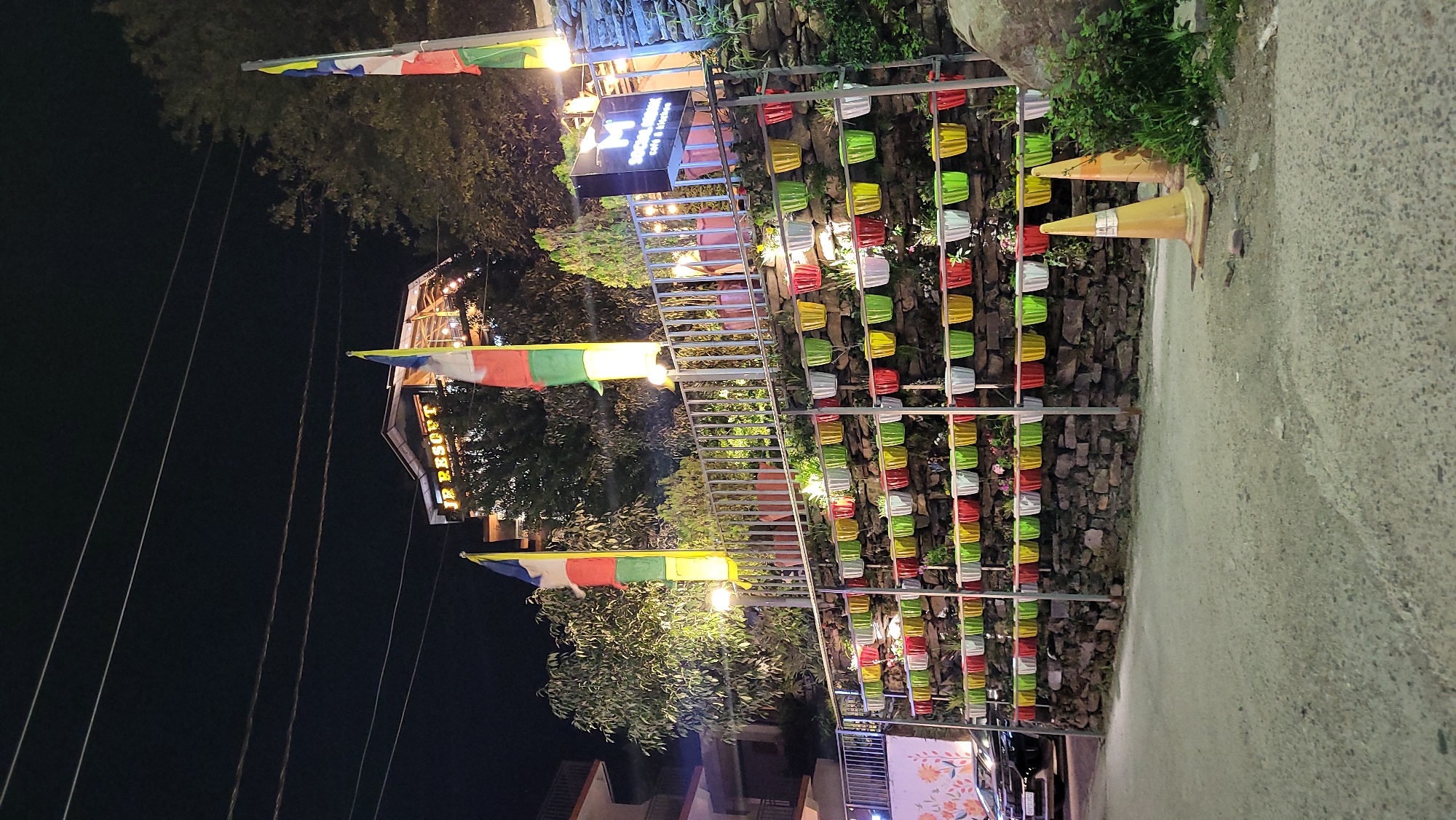 The image depicts a street scene at night in Haini, India. On the right side of the frame, a stone wall is adorned with multiple rows of colorful flower pots. In front of the wall, two orange traffic cones are positioned on the edge of the road. A metal gate-like structure stands in front of the stone wall.
On the left side of the frame, Tibetan prayer flags are visible, strung across the street. A building is visible, with the words "Social Hour Cafe & Restro" displayed on a sign. Further back, there is a building labeled "JR Resort." The street is illuminated by streetlights. No people are visible in the image.
