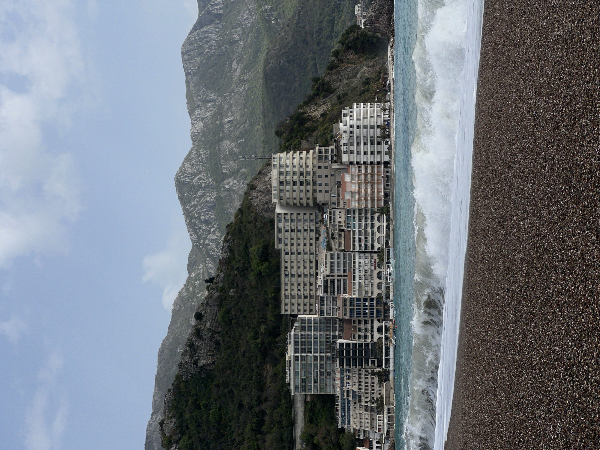 The image depicts the city of Boreti, Montenegro, nestled between mountains and the Adriatic Sea. A line of multi-story hotels and apartments hugs the coast, facing the beach. Waves crash onto a pebble beach. The buildings are a mix of modern and older styles. Mountainous terrain rises steeply behind the buildings. The sky is overcast. The beach is dark grey/brown. There are no people visible in the image.