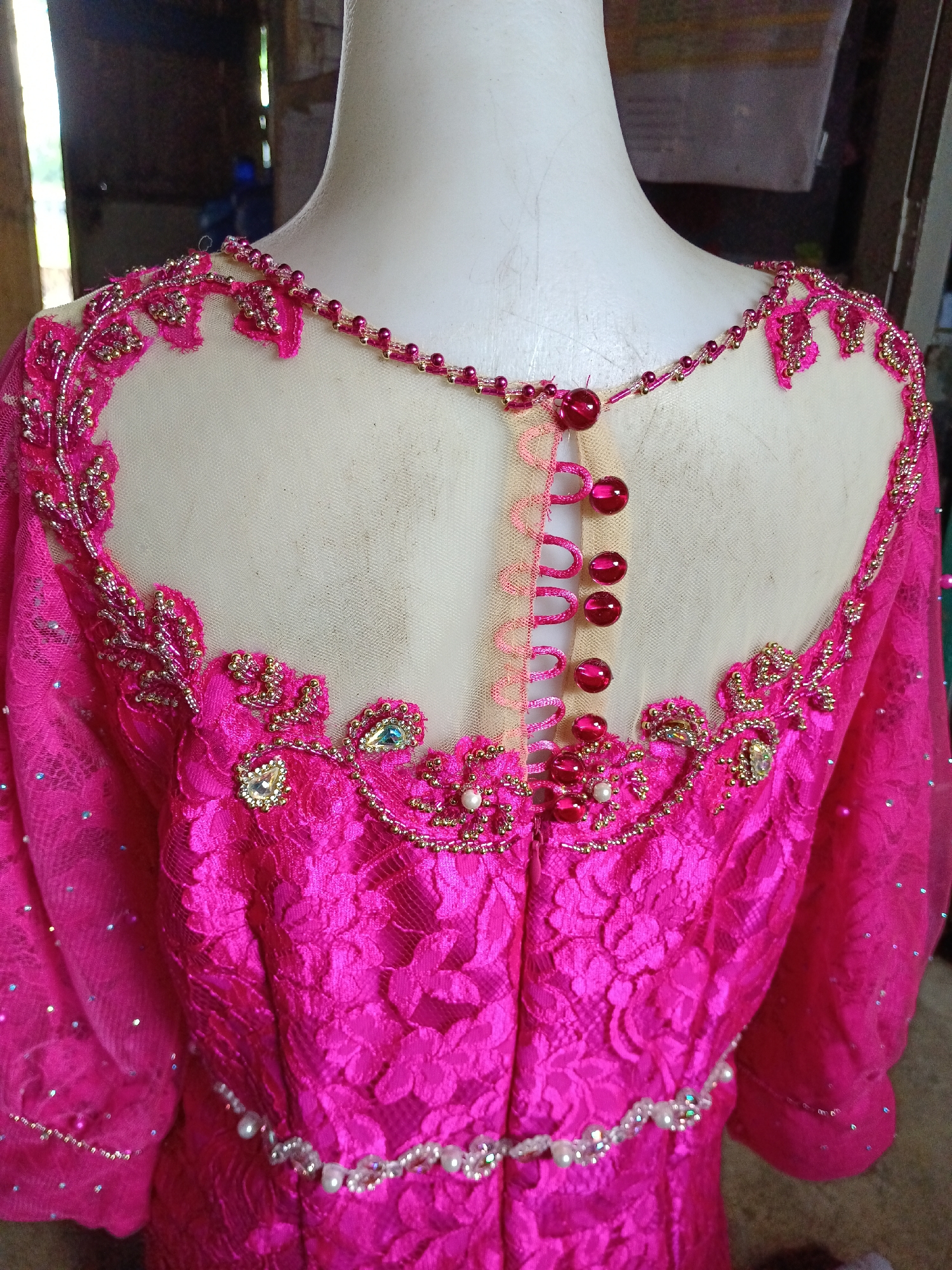 A bright pink, lace dress adorned with beads, pearls, and sequins is displayed on a white mannequin. The dress features a sheer neckline with intricate beaded embellishments around the collar and shoulders. A row of five pink, glossy buttons is visible down the back, fastened with delicate pink loops. The sleeves are sheer and also decorated with small scattered sequins. A beaded belt with pearls and crystals cinches the waist. The background is blurred, showing a rustic, wooden structure with hints of greenery, suggesting an outdoor or workshop setting. The image was taken in Aek Kanopan, Indonesia, likely within a tailor's shop or a personal residence where the dress is being showcased or worked on.