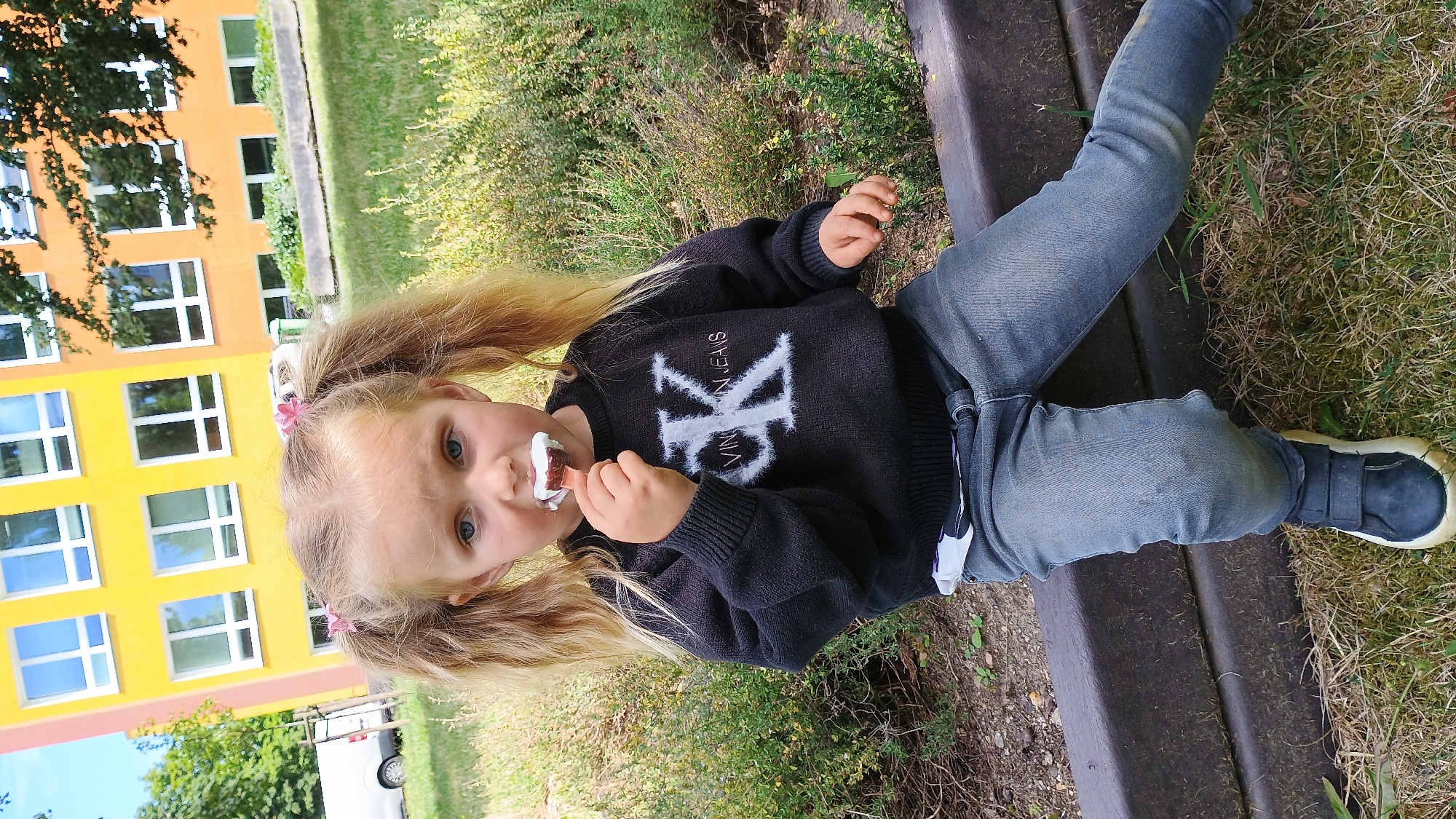 A young blonde girl with pigtails is eating an ice cream bar. She is wearing a black sweater with a white "K" logo and blue jeans, and dark blue sneakers. She is sitting on a dark brown bench in front of a multi-story yellow and orange building. The building has many windows and appears to be a school. The girl is outdoors on a sunny day. The ground is covered with grass and some scattered gravel. In the background, there are some trees and a white van parked on a street. The location is Sokolov, Czechia.