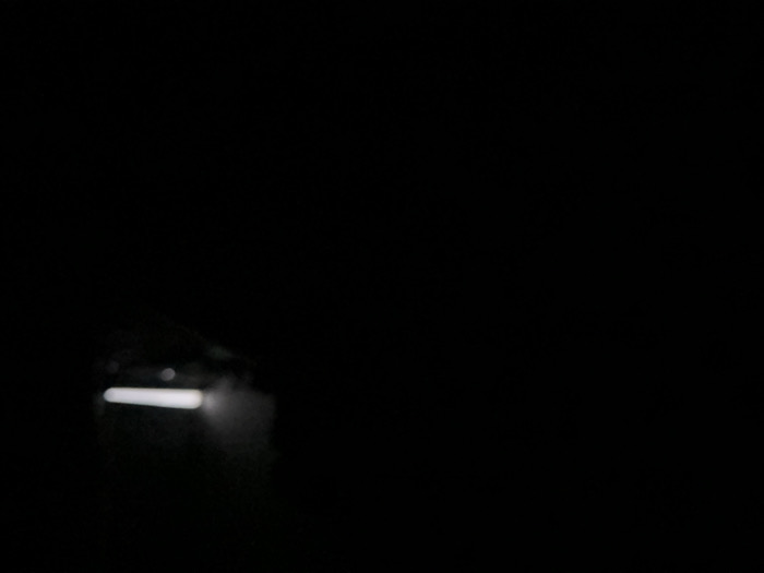 In the dark, a bright, horizontal light source illuminates a small area. The light appears diffused, surrounded by dark, indistinct shapes. The overall impression is of a focused point of light breaking through the surrounding darkness, possibly a flashlight in a dark room.