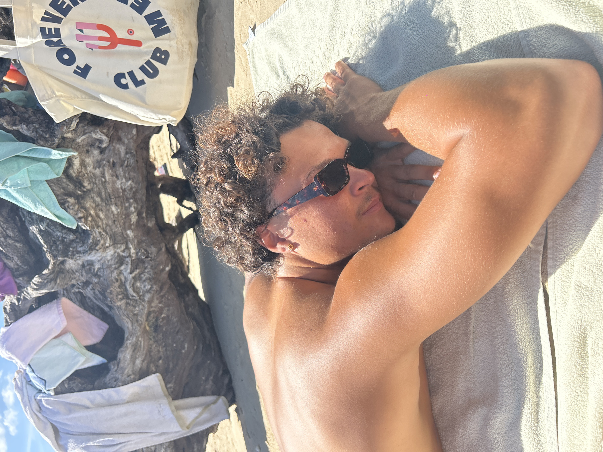 A young individual with tanned skin, curly brown hair, and wearing tortoiseshell-patterned sunglasses and a gold stud earring in the left ear is depicted lying face down, partially on their left side, on a light-colored terry cloth towel spread on a sandy beach. The individual is shirtless, with their head resting on their forearms. In the background, a large, gnarled piece of driftwood or a tree trunk occupies the mid-ground. Several pieces of clothing or small towels in various light shades (white, light blue, light pink, light green) are draped over the driftwood. To the right of the driftwood, a cream-colored canvas tote bag is visible, featuring "FOREVER CLUB MED" in dark blue text encircling a stylized red and pink trident logo. The scene is illuminated by bright daylight under a partially cloudy blue sky. The location is a beach in Sainte-Anne, France.