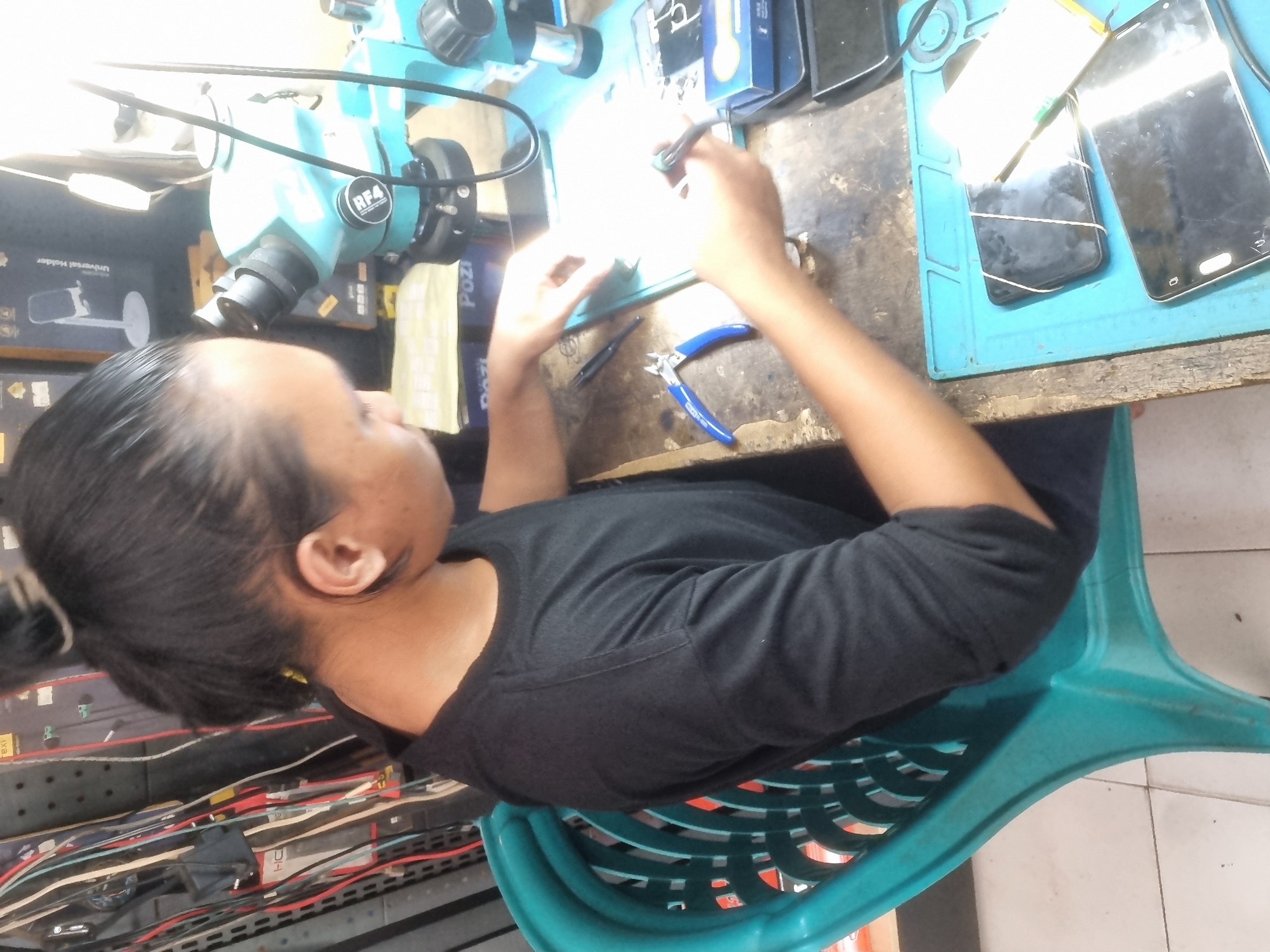 A person with dark hair pulled into a bun is seated on a turquoise plastic chair, facing a workbench. They are wearing a black long-sleeved shirt and are looking down at the workbench, which is cluttered with electronic repair tools and parts. In front of them is a blue mat with several mobile phones, some of which are held together with rubber bands. A microscope with a blue base is positioned on the workbench, along with various small tools like pliers and tweezers. Wires are visible hanging on a rack in the background. The scene appears to be a repair shop, potentially in Probolinggo, Indonesia, given the context.