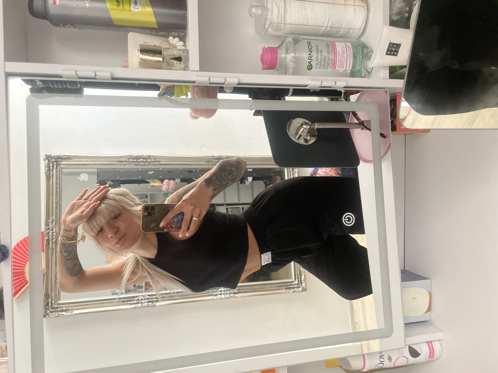 A woman with blonde hair and tattoos is taking a selfie in a mirror. She is wearing a black crop top and black sweatpants. The mirror is framed in silver and is mounted on a wall in a bathroom or dressing area, with shelves visible above and below. The shelves contain various toiletries and cosmetic products. The location is likely a private residence, but there is no information to suggest that this image was taken in Tilburg, Netherlands.