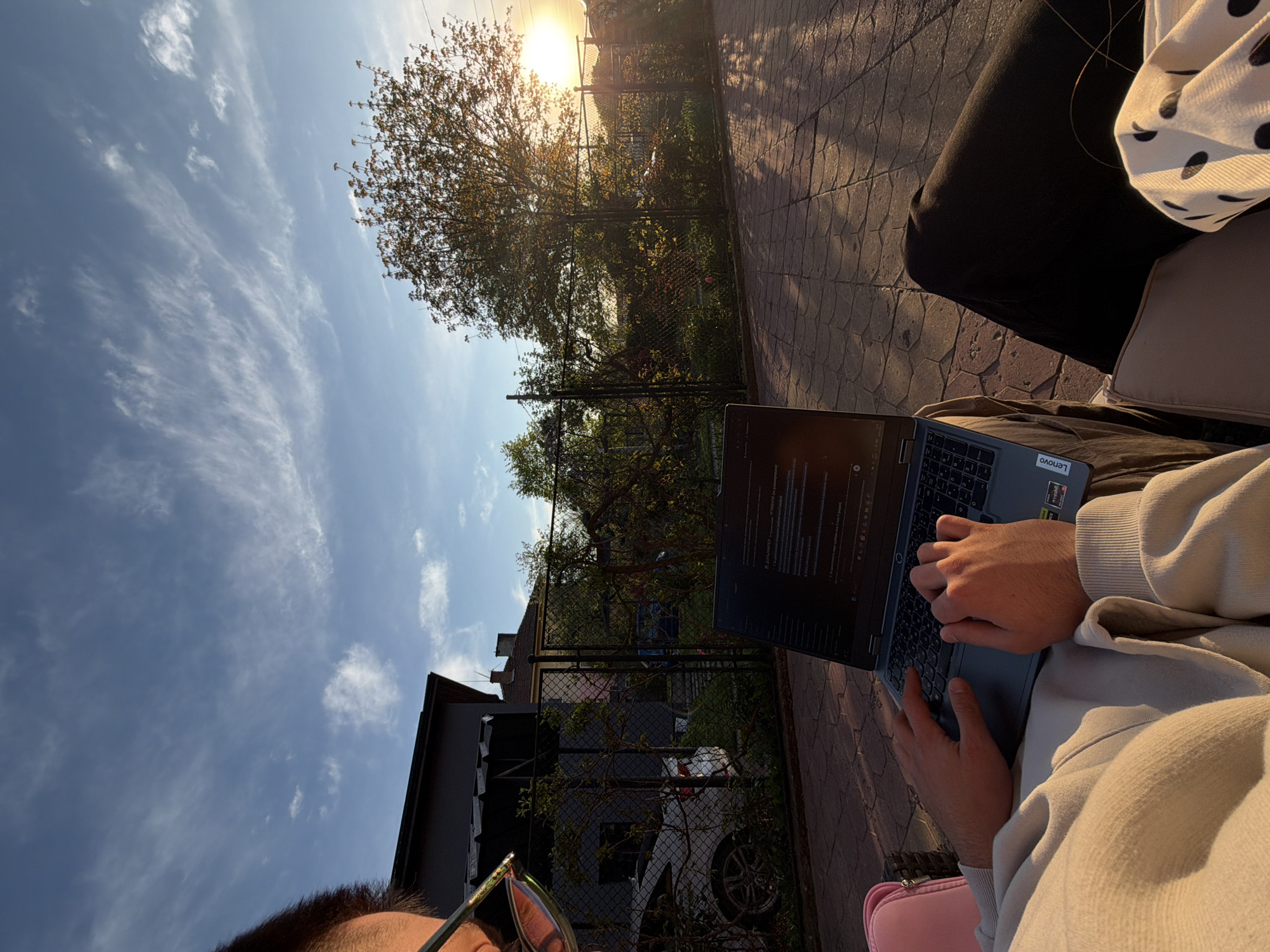 The image captures an individual using a Lenovo laptop outdoors. The person is sitting on a patio with a brick or tile floor. The person is wearing a light-colored hoodie. Visible outside the patio area is a tree, a fence, a house, and a car. The sky is blue with some clouds, and the sun is shining brightly. The picture was taken outdoors in what seems to be Buhovo, Bulgaria.