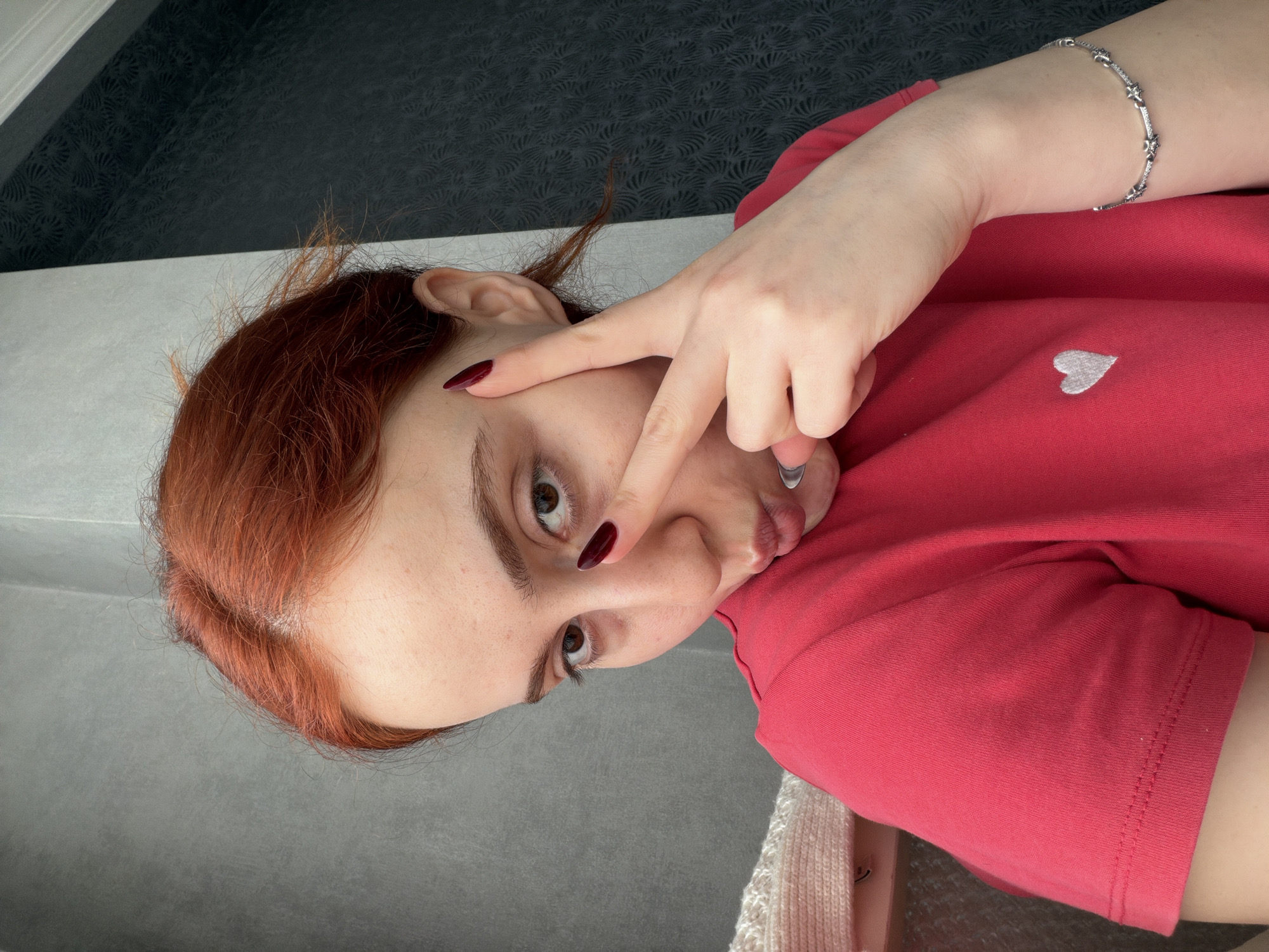 A young woman with reddish-brown hair and fair skin is captured in a close-up, angled shot. She is lying down, looking towards the camera with her dark brown eyes. Her left hand is raised, with her index and middle fingers making a "V" shape and resting near her left eye. She has dark red nail polish on her fingernails. Her lips are slightly pursed in a playful or casual expression.
She is wearing a coral-colored or bright red t-shirt, which features a small, light gray embroidered heart on the left chest area. On her right wrist, she wears a slender silver bracelet adorned with small, star-like or bow-shaped elements.
The background is simple and divided horizontally. The lower section consists of a plain light gray surface, while the upper section is a darker gray or black patterned surface, possibly wallpaper or fabric with a subtle repeating design. No specific outdoor location cues are visible. There is no visible text in the image. The lighting appears even, suggesting an indoor setting.