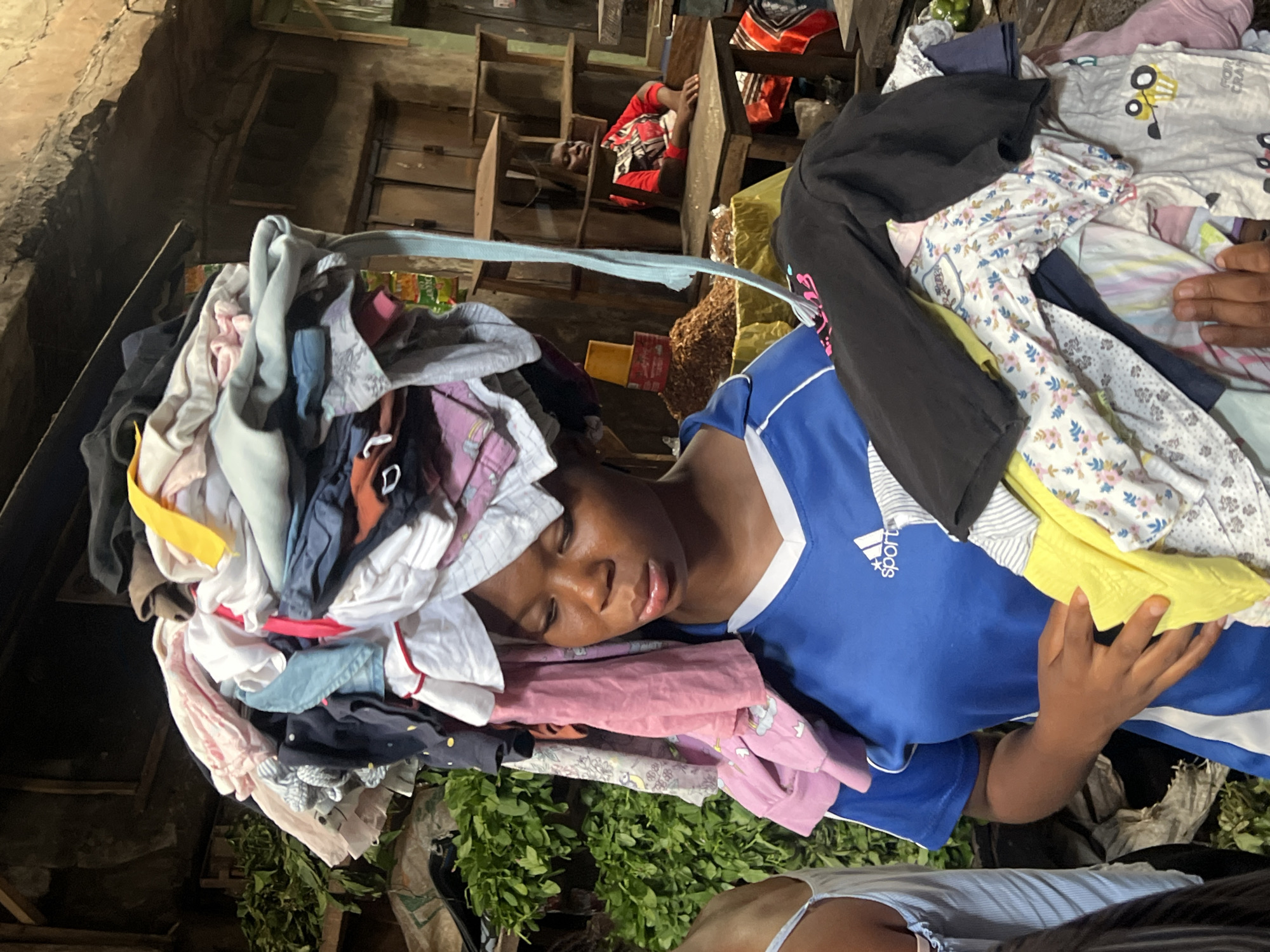 The image depicts a young person sleeping amidst a large pile of colorful clothing. The individual, dressed in a blue and white athletic shirt, is lying on their side with their head resting on a heap of laundry, which includes various garments such as t-shirts, pants, and pajamas in an assortment of colors and patterns. The pile of clothes is substantial, creating a makeshift bed for the sleeping person.
The setting appears to be indoors, possibly a market stall or a storage area, given the rustic wooden shelves and stacked crates visible in the background. The overall environment seems somewhat cluttered and utilitarian. Green leafy branches are visible at the bottom of the frame, suggesting the presence of plants or perhaps goods being sold.
A person in a red and black patterned shirt is also visible in the background, resting on a wooden surface, suggesting that this is a place where people may rest among the goods. The lighting suggests it could be daytime, though the interior setting makes it difficult to ascertain the exact time of day or weather conditions.
The expression on the sleeping person's face is relaxed, indicating peaceful rest. The focus of the image is on the sleeping individual and the surrounding clothing, highlighting a moment of repose within a busy or perhaps working environment. Some text is visible on the blue shirt, including the Adidas logo and the word "sports". Another garment in the pile has a visible cartoon character. The image has a vertical orientation.