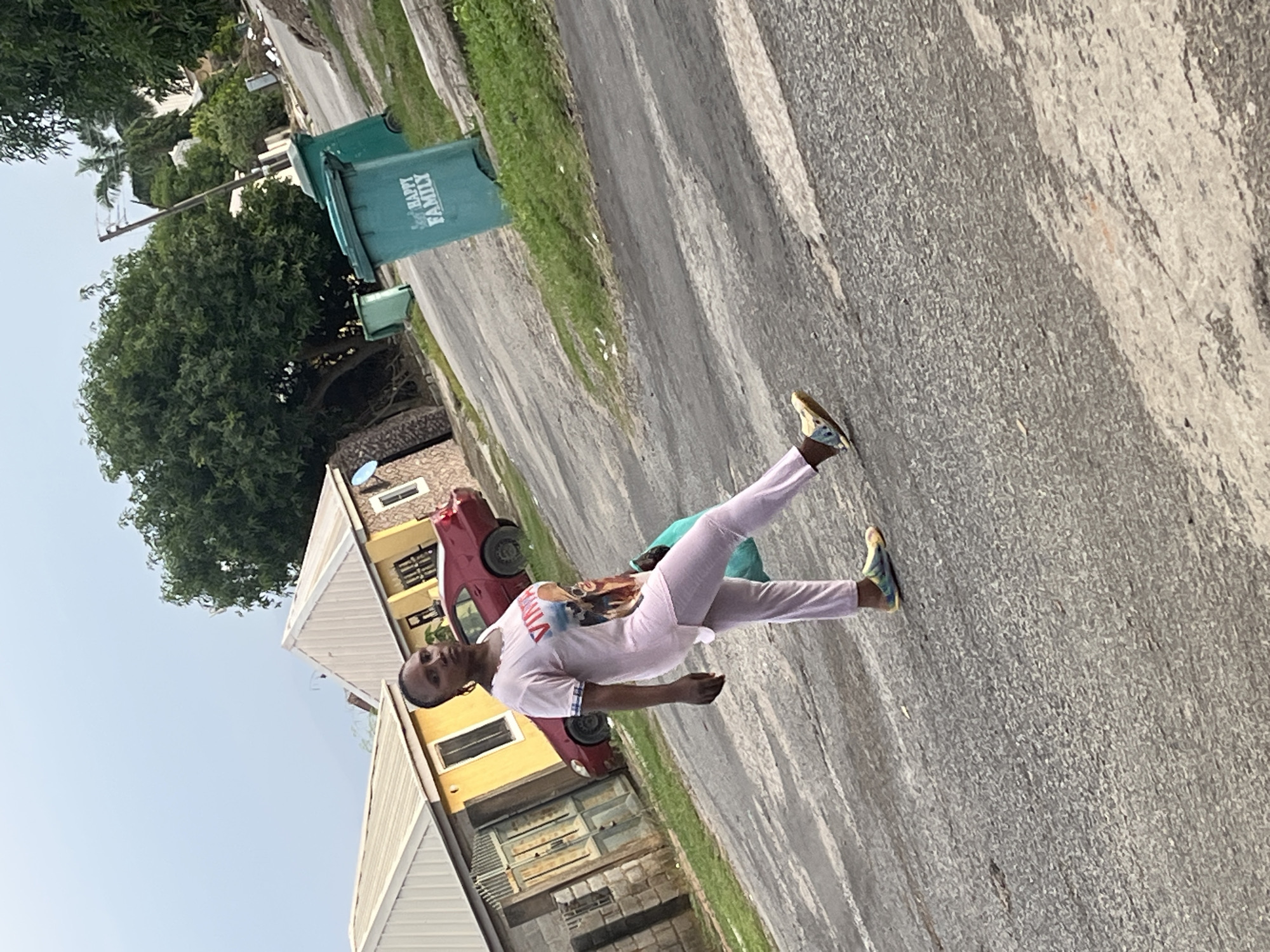 The image depicts a street scene in Abuja, Nigeria, with a woman walking on an asphalt road. She is wearing a light pink t-shirt with a graphic print and matching pink trousers. On her feet are colorful sandals. She is carrying a light blue bag in her left hand.
In the background, there are residential buildings with yellow exteriors and white roofs. A red car is parked in front of one of the houses. Several green trash bins are visible along the side of the road, with one prominently displaying the words "Happy Family" in white lettering. The environment appears to be a typical residential street with vegetation and trees. The sky is overcast, suggesting a cloudy day.