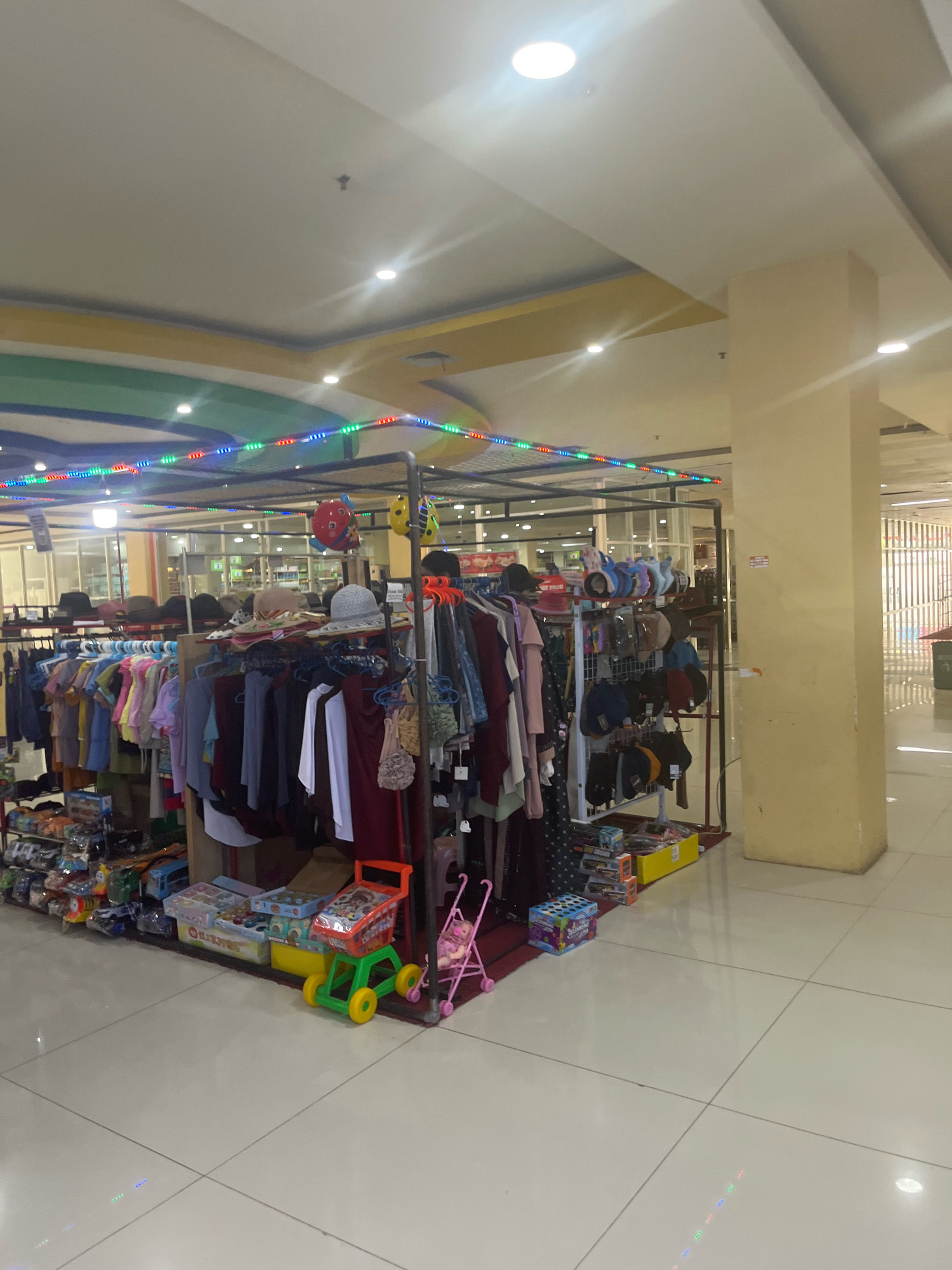 The image captures a retail stall within a shopping mall in Sengkang, Indonesia. The stall is densely packed with various items, primarily clothing and accessories displayed on racks and shelves. A rack in the foreground is filled with a variety of colorful garments, including shirts, dresses, and skirts, all hung on hangers. Above the clothing, several hats are displayed, including fedoras, wide-brimmed hats, and baseball caps. Some small toys, including a toy shopping cart and a doll stroller, are placed on the floor in front of the stall. The stall is illuminated by overhead fluorescent lights and decorated with a string of colorful LED lights that drape across the top of the structure. The background of the image shows more of the shopping mall's interior, including tiled floors, structural columns, and other retail spaces visible through glass partitions. The overall impression is one of a busy marketplace stall, offering a diverse range of goods to shoppers.