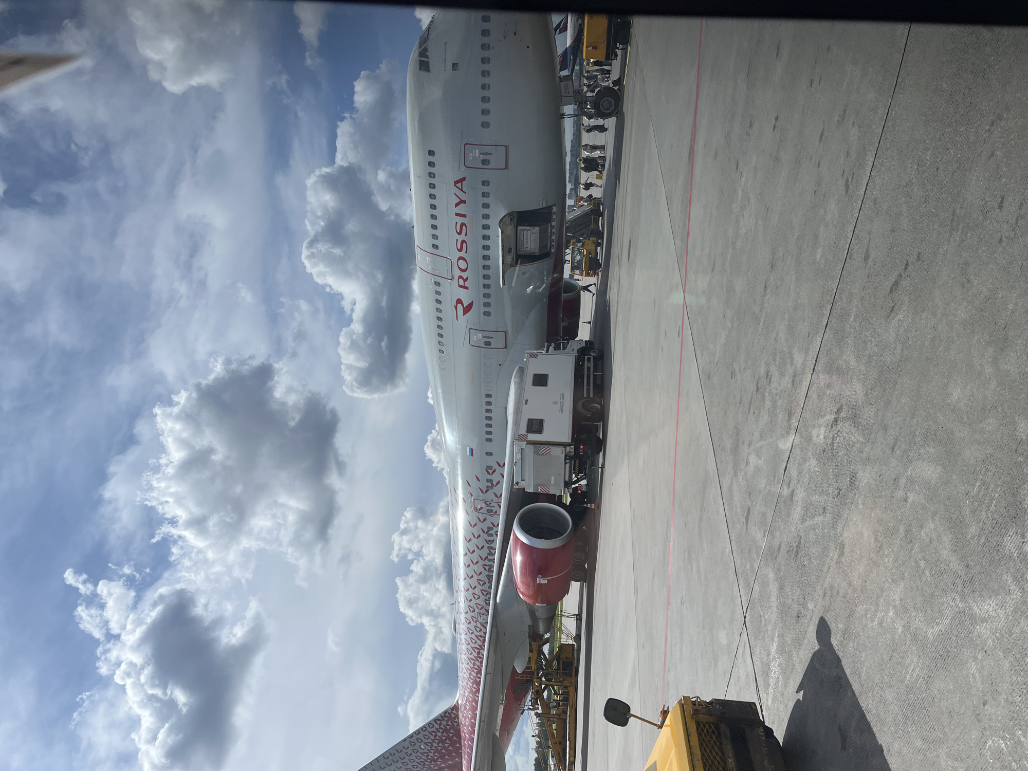 An exterior shot of a Rossiya Airlines Boeing 747 aircraft parked on the tarmac at Khimki's Sheremetyevo International Airport. The aircraft, predominantly white with red accents and a patterned tail, is situated under a partly cloudy sky. Ground crew and passengers are visible near the fuselage, some boarding or disembarking via a mobile staircase. Various airport vehicles, including a baggage tug and a service truck, are positioned around the plane. The tarmac is paved and marked with lines, and a yellow vehicle is partially visible in the foreground.
