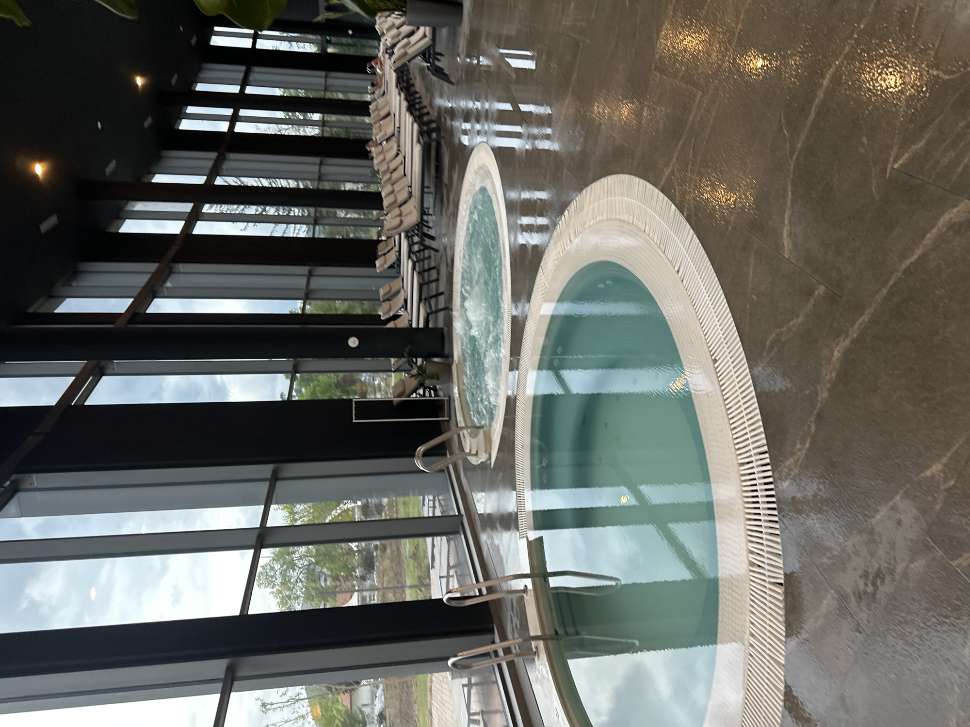 An indoor spa or wellness area is depicted, featuring two circular hot tubs with light-colored, ridged surrounds. The larger hot tub is in the foreground, and a slightly smaller one is positioned further back, both containing clear, blue-green water. The floor is made of a dark, glossy, possibly marbled tile, appearing wet and highly reflective, mirroring the warm glow of several recessed ceiling lights.
To the left, a wall of tall, dark-framed, floor-to-ceiling windows offers a panoramic view of the outdoors. Through these windows, a cloudy sky and green trees are visible, suggesting an overcast day. Along the far wall, a row of light-colored lounge chairs or recliners with dark frames is arranged, indicating a relaxation zone.
The ceiling is dark, with circular recessed lights. Metal handrails are visible, leading into the hot tubs. A large green plant is partially visible in the upper left corner, adding a touch of nature to the indoor setting. The scene is tranquil and modern, suggesting a luxurious environment designed for relaxation. There are no clear human figures engaged in specific activities, and no visible text is present. The location context is Kazanlak, Bulgaria.