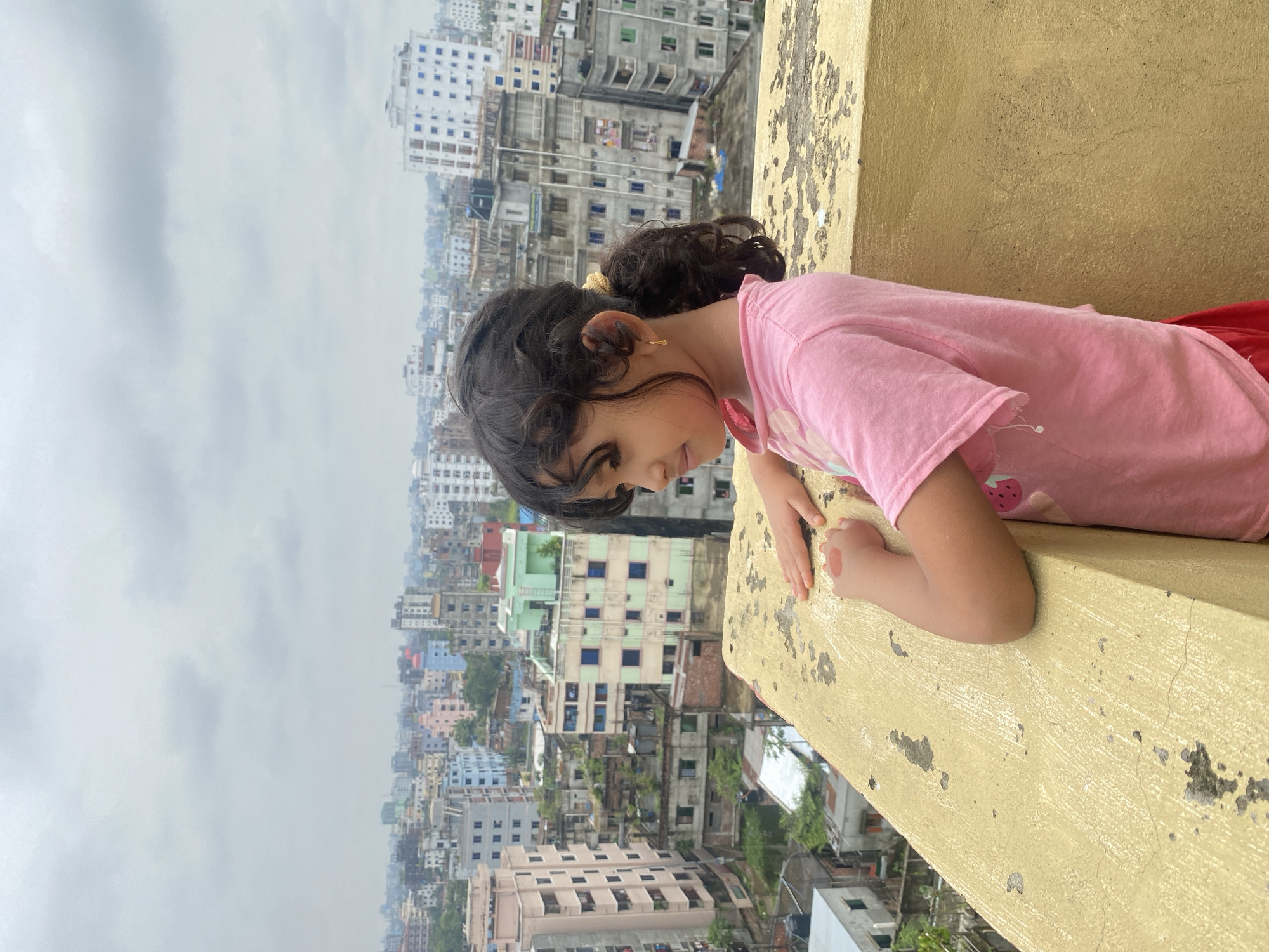 A young girl stands on a yellow, weathered rooftop overlooking a densely populated cityscape of Dhaka, Bangladesh. The sky is overcast with clouds. The girl, with dark, curly hair pulled into a ponytail, wears a pink t-shirt with a strawberry graphic and red bottoms. Her right hand rests on the rooftop ledge, and her left arm is bent, with her hand near her chest. She is looking off to her left, with a pensive expression. The buildings in the background are a mix of modern and older structures, varying in color and height, with some showing signs of wear and tear. The overall impression is of an urban environment under a cloudy sky.
