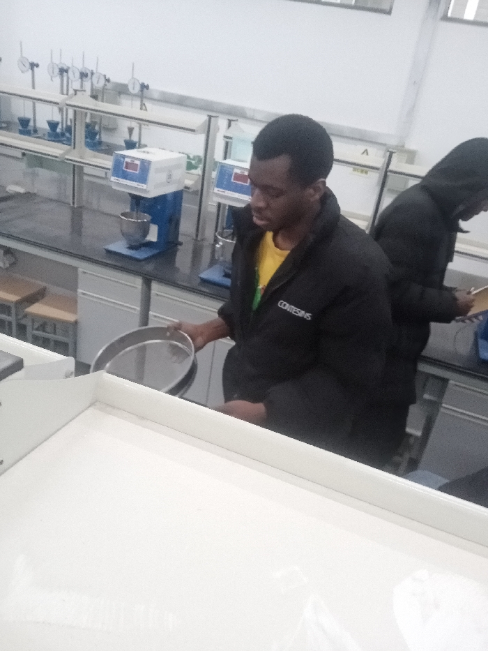 In a lab setting, a man in a black jacket and yellow shirt holds a sieve. Another individual, also in black, stands behind him, possibly taking notes. Lab equipment is visible in the background, including machines with bowls and various gauges. The scene suggests a scientific experiment or analysis is in progress. A large white surface is in the foreground.