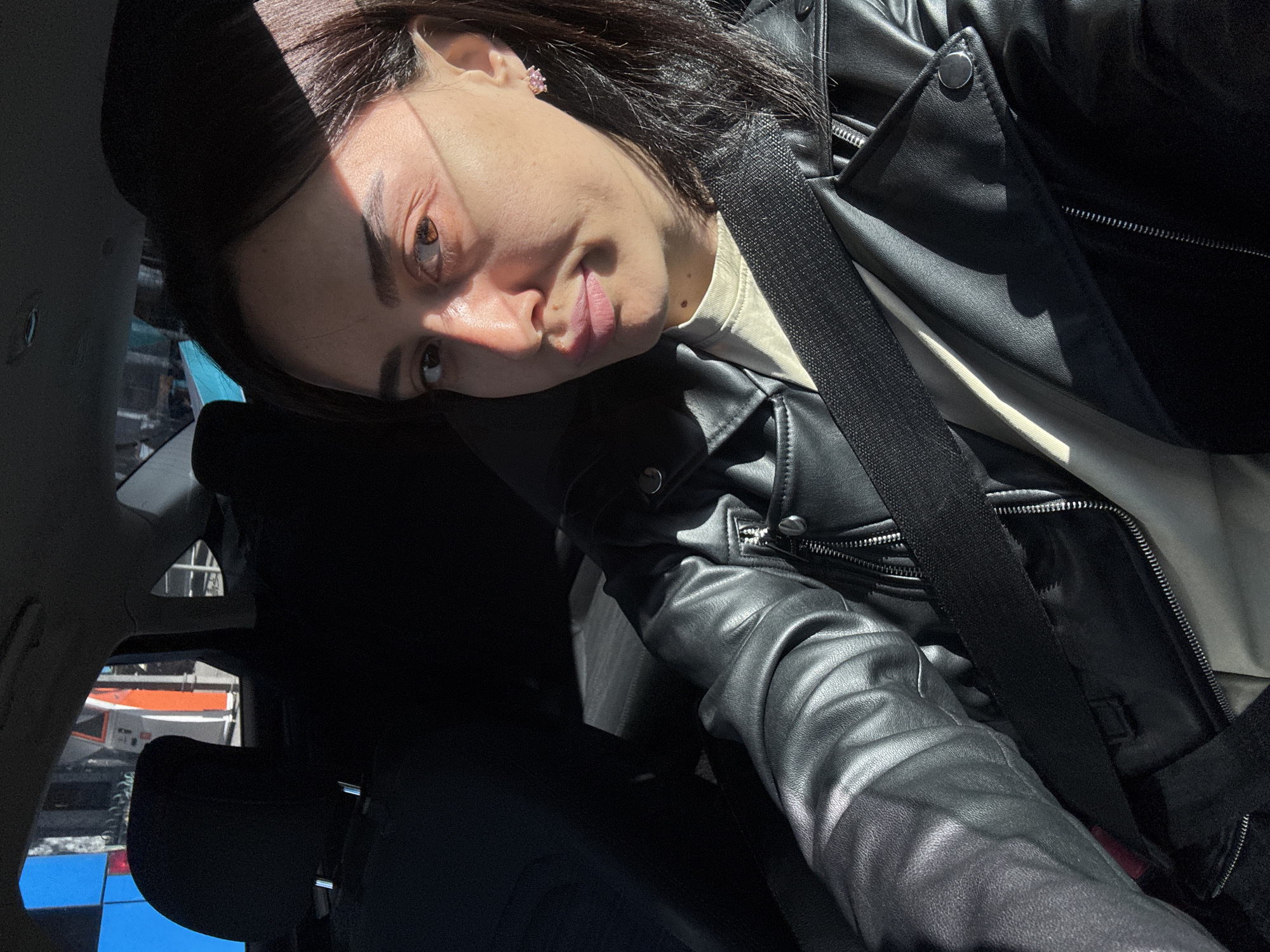 A medium shot, taken inside a vehicle, shows a young woman with dark hair, brown eyes, and fair skin. She is wearing a black leather jacket over a light-colored shirt and a black seatbelt. Her head is resting on the seat back. Light enters the vehicle and casts shadows across her face. Through the car window, scaffolding and an orange and blue lift truck are visible, indicating an urban construction site. The photograph was possibly taken in the city of Tbilisi, Georgia.